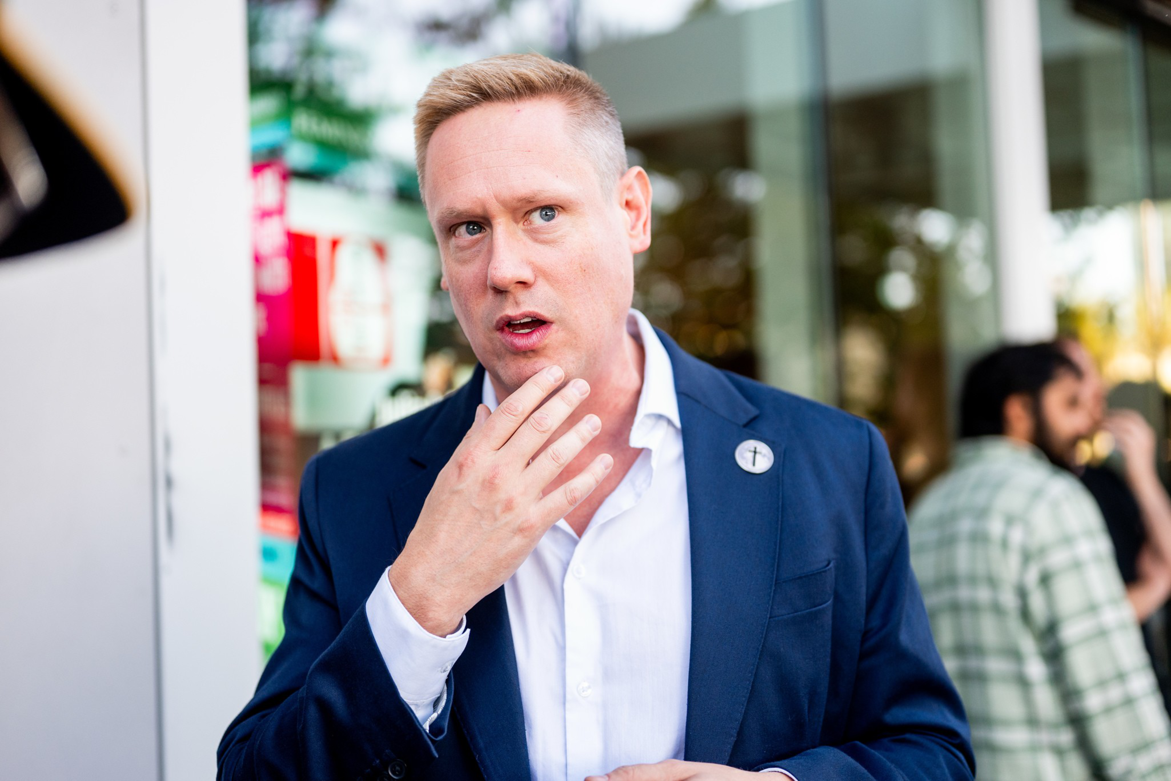 A man in a navy blazer and white shirt touches his chin thoughtfully, standing outside near a glass window with blurred people in the background.