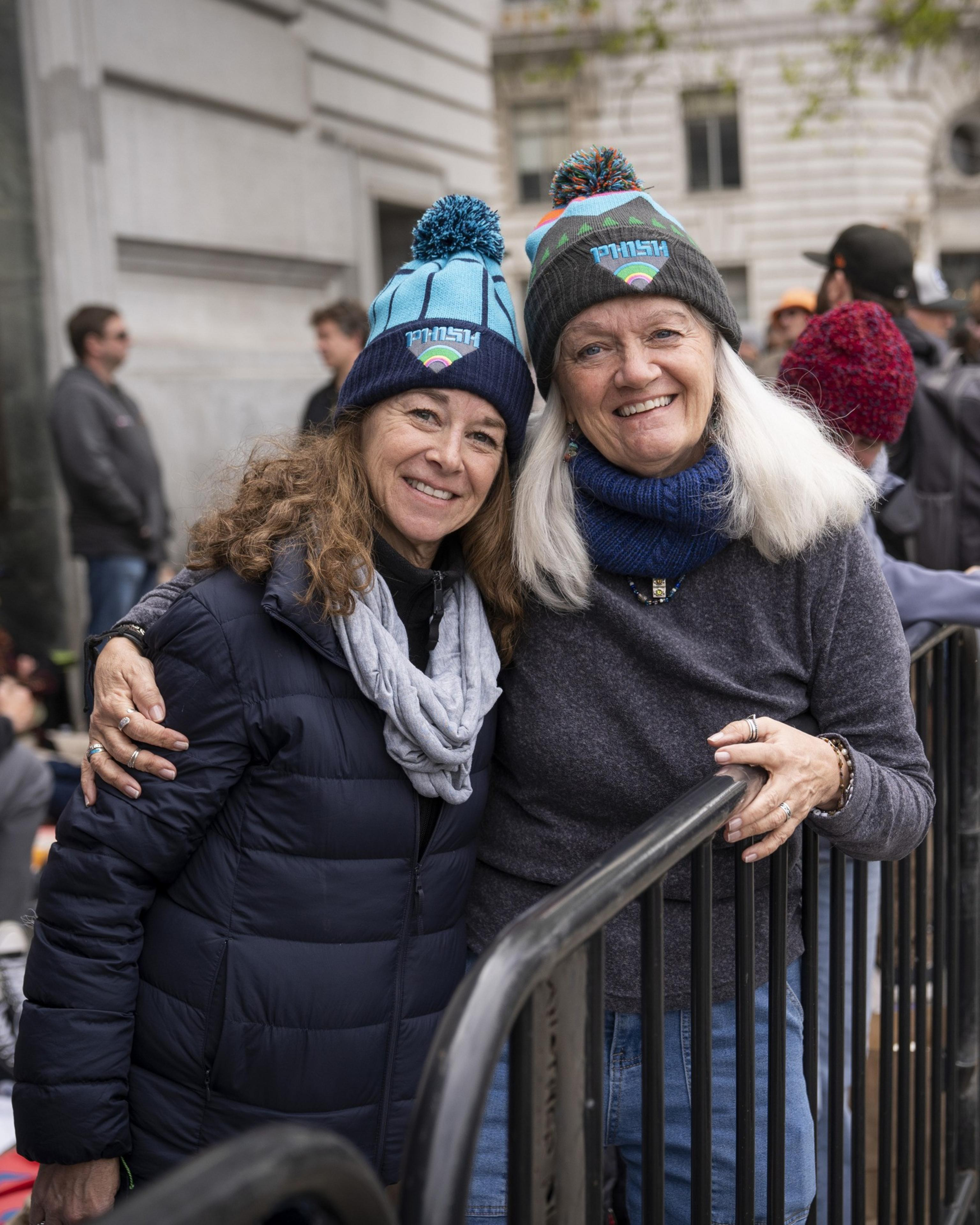 Two smiling women are wearing winter hats and jackets, with their arms around each other, standing behind a railing at an outdoor gathering.