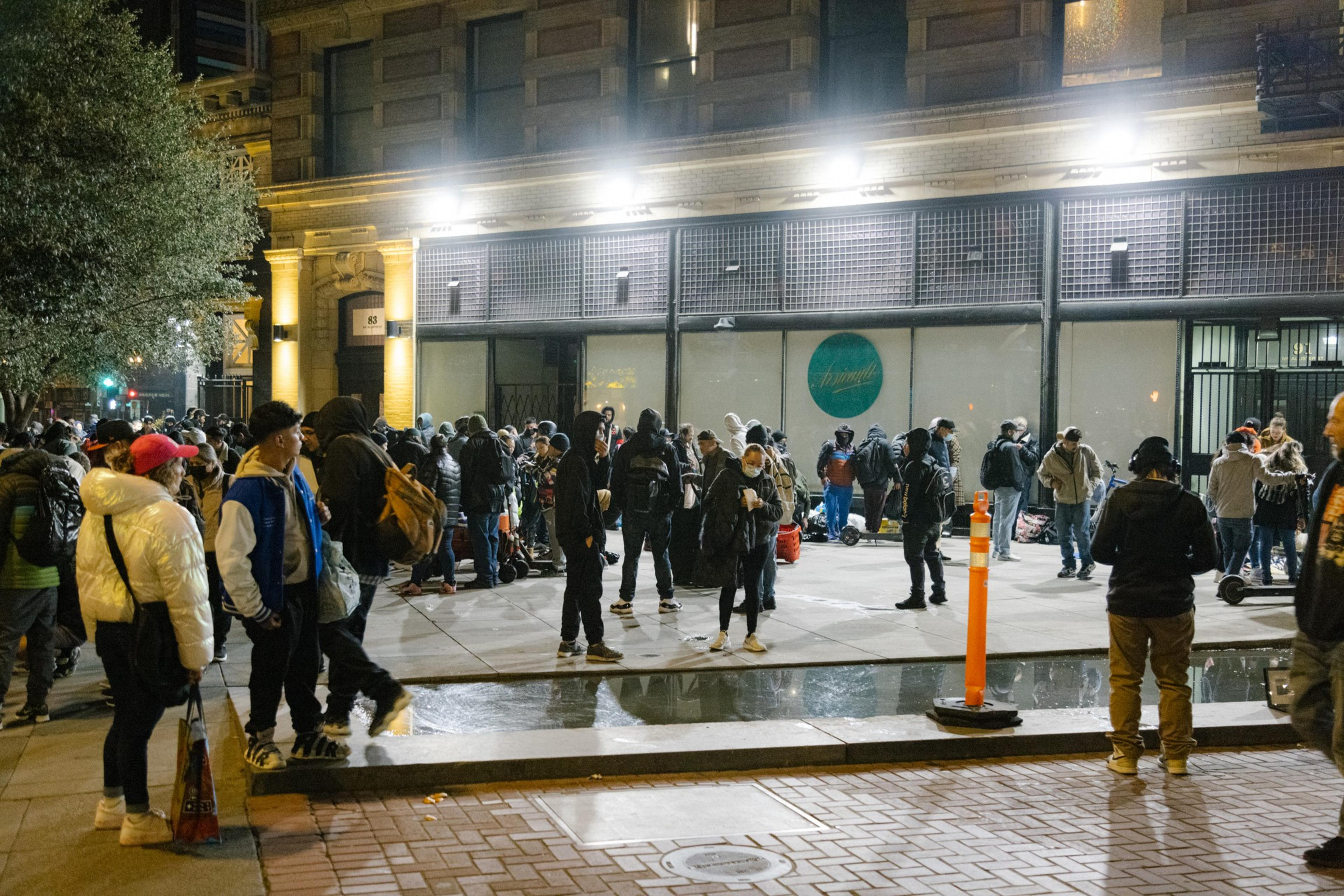 A large group of people gathered at night on an urban street, some with bags, near buildings with lit windows.