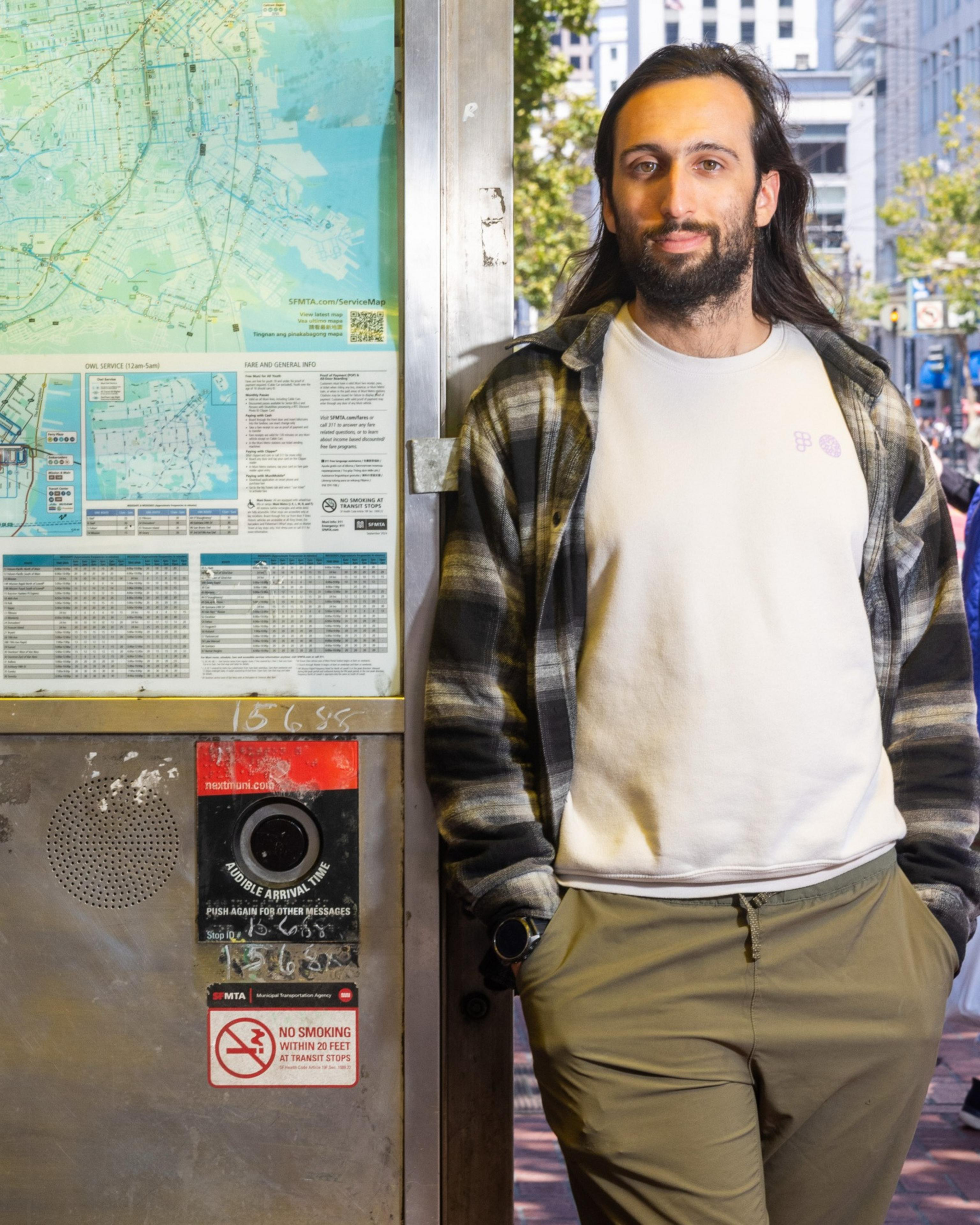 A person with long hair and a beard is leaning against a public transit map display. They wear a plaid shirt, white t-shirt, and khaki pants, with hands in pockets.