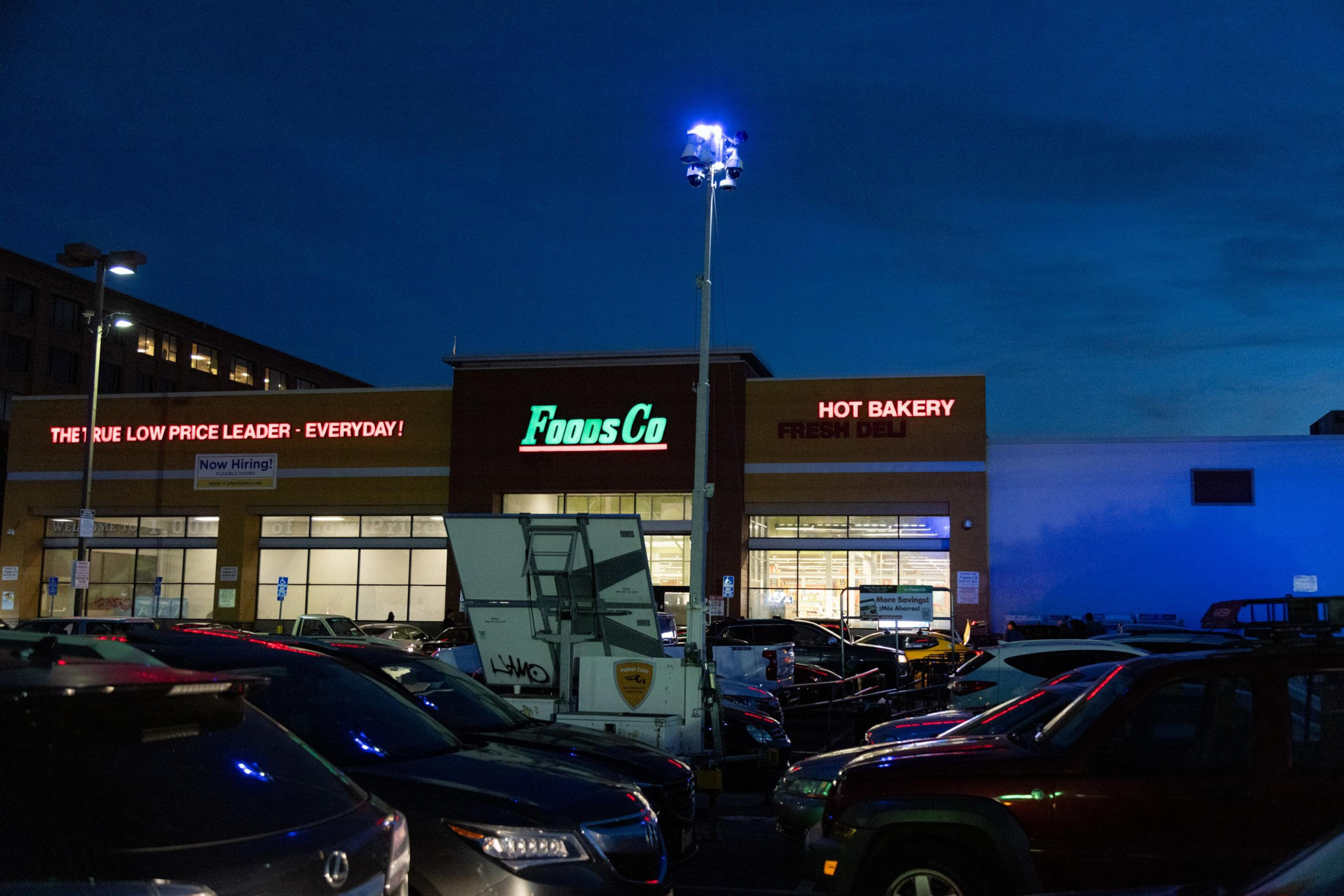A security camera tower sits in the parking lot of Foods Co. at night with cars surrounding it. 