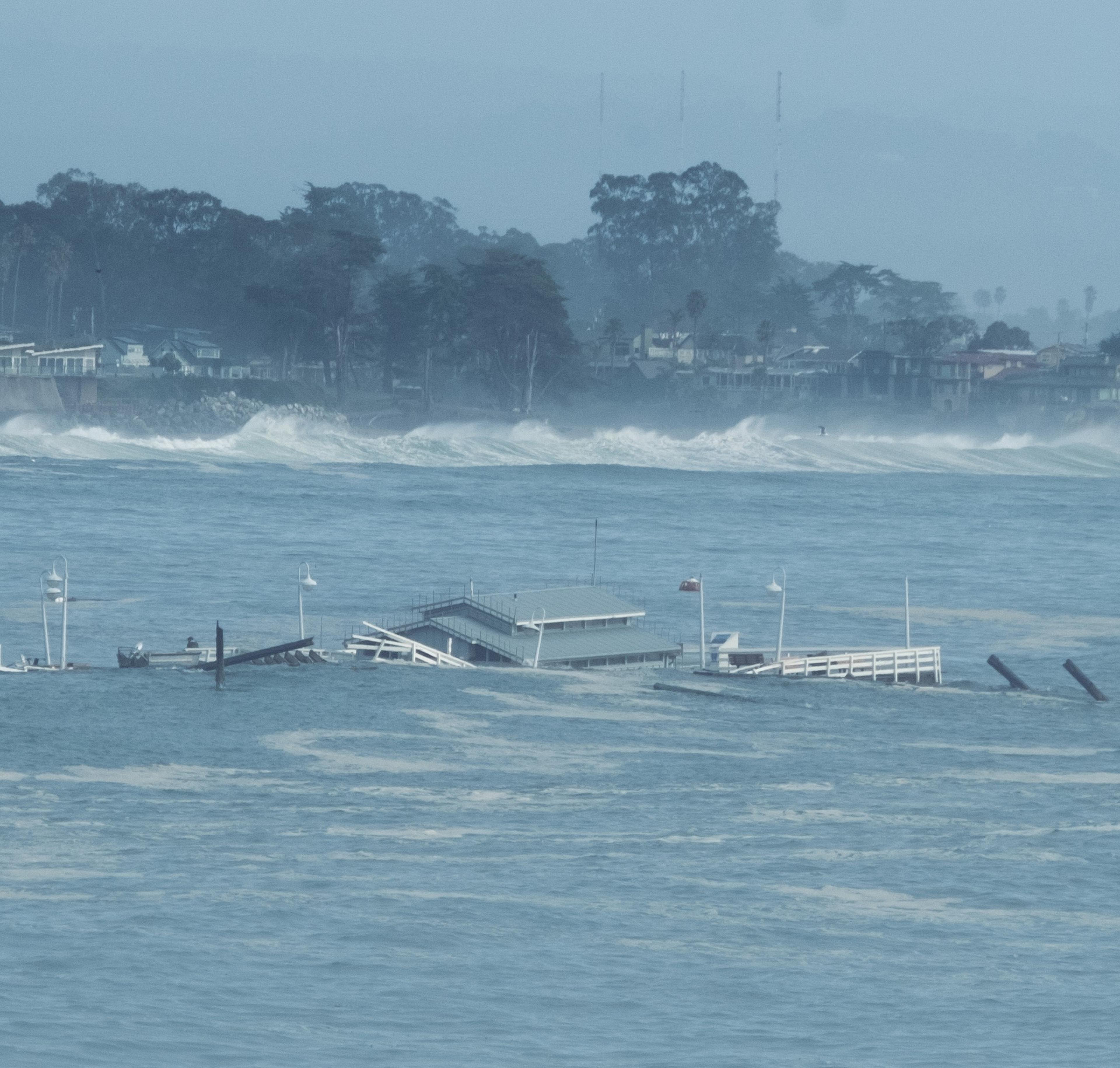 A partially submerged pier with scattered debris is visible in rough ocean waters, with waves crashing closer to a shoreline lined with trees and buildings.