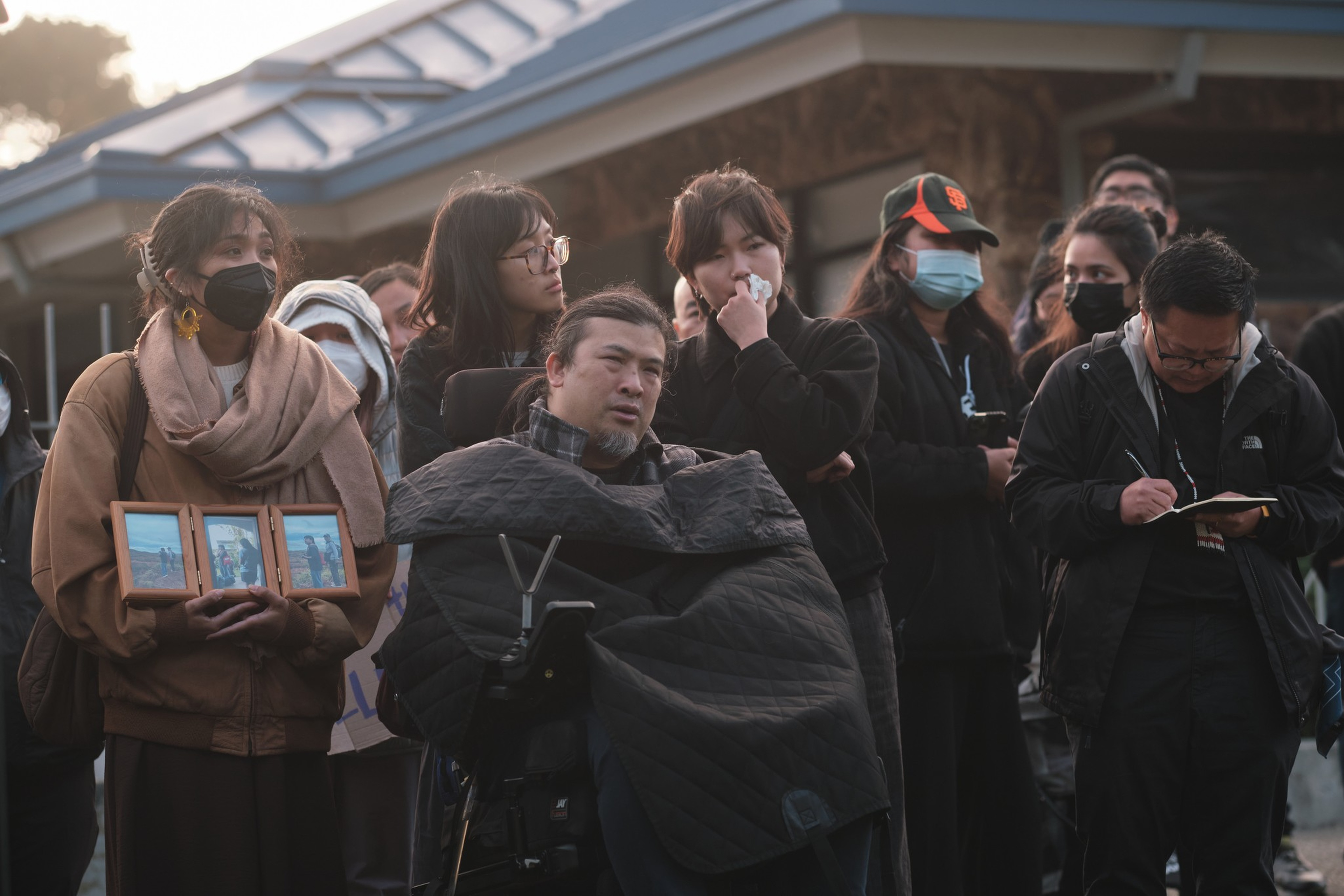 A group of solemn people stand close together outdoors, one holding framed photos, another using a wheelchair wrapped in a black blanket.