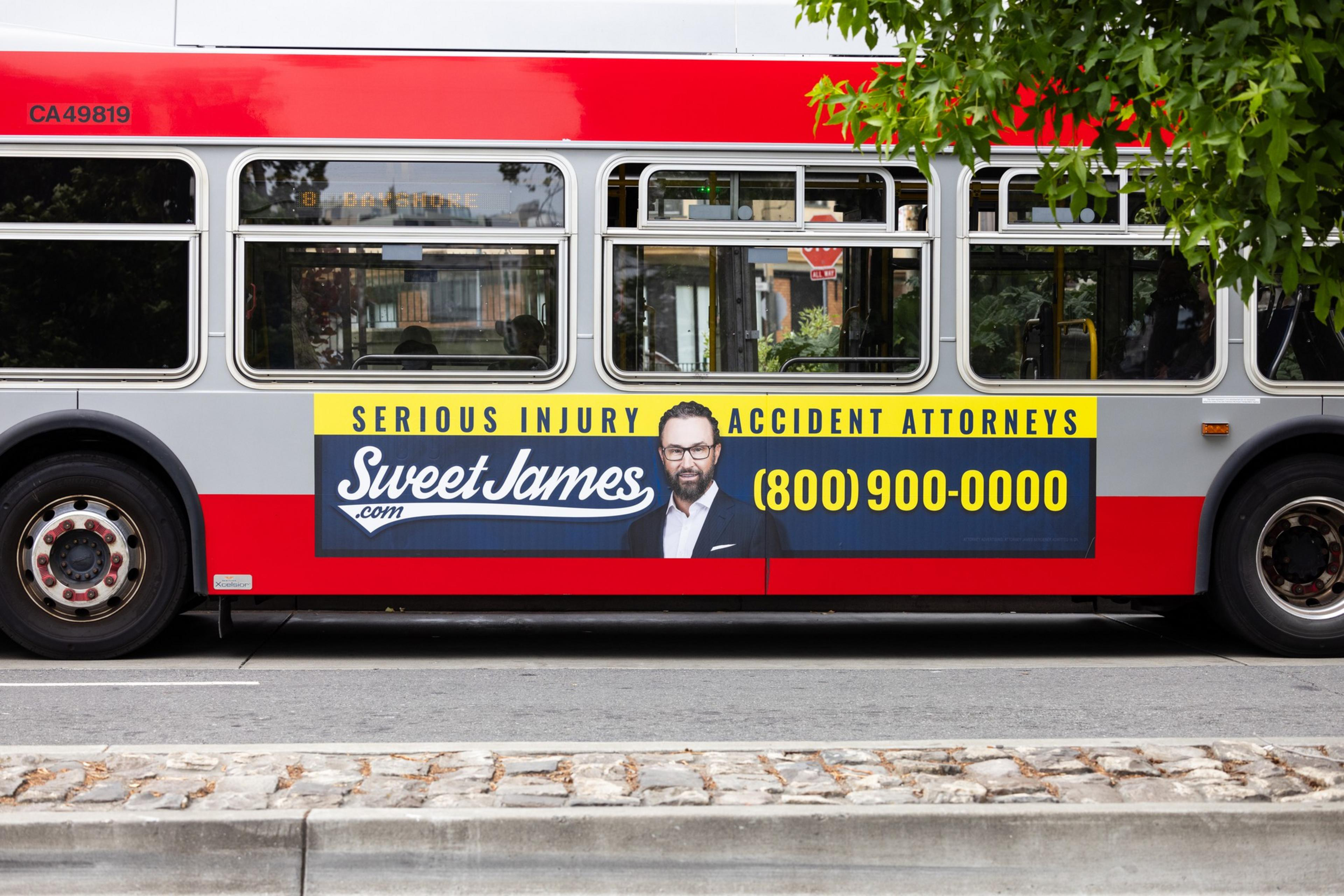 The side of a red and silver bus displays an ad for "Sweet James Serious Injury Accident Attorneys" with a picture of a man, website, and phone number (800) 900-0000.