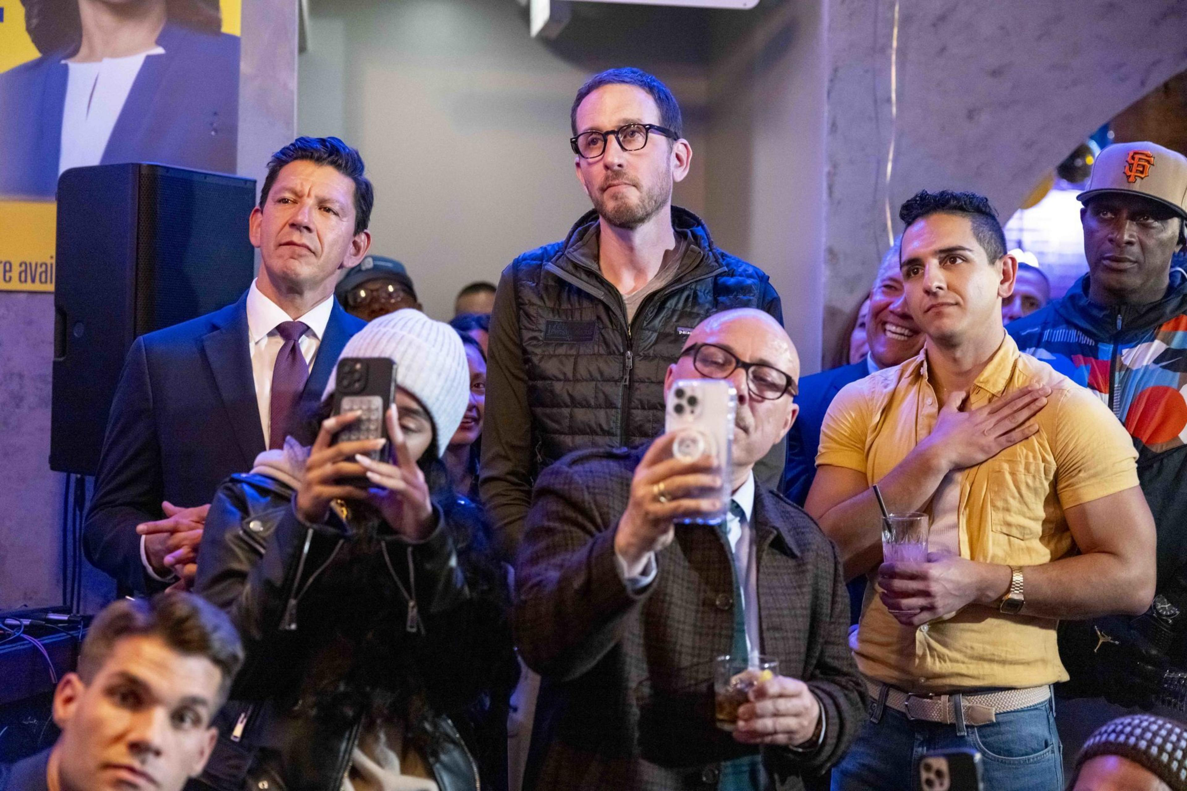 A group of people are gathered indoors, some taking photos with phones. They appear attentive and engaged, with a mix of casual and formal attire.