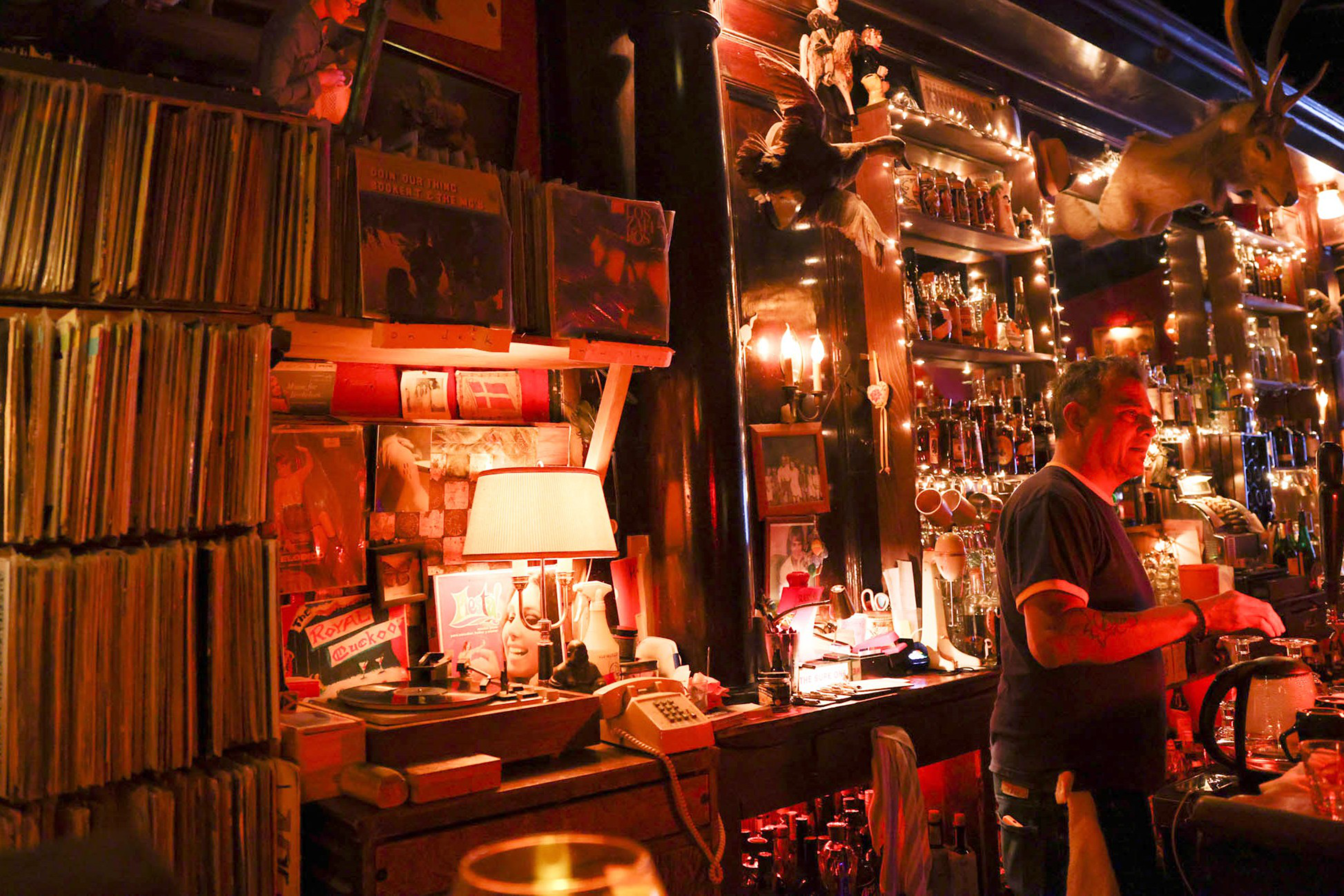 A warmly lit bar with shelves of liquor, a man behind the counter, vinyl records, a record player, a lamp, and a mounted deer head on the wall.