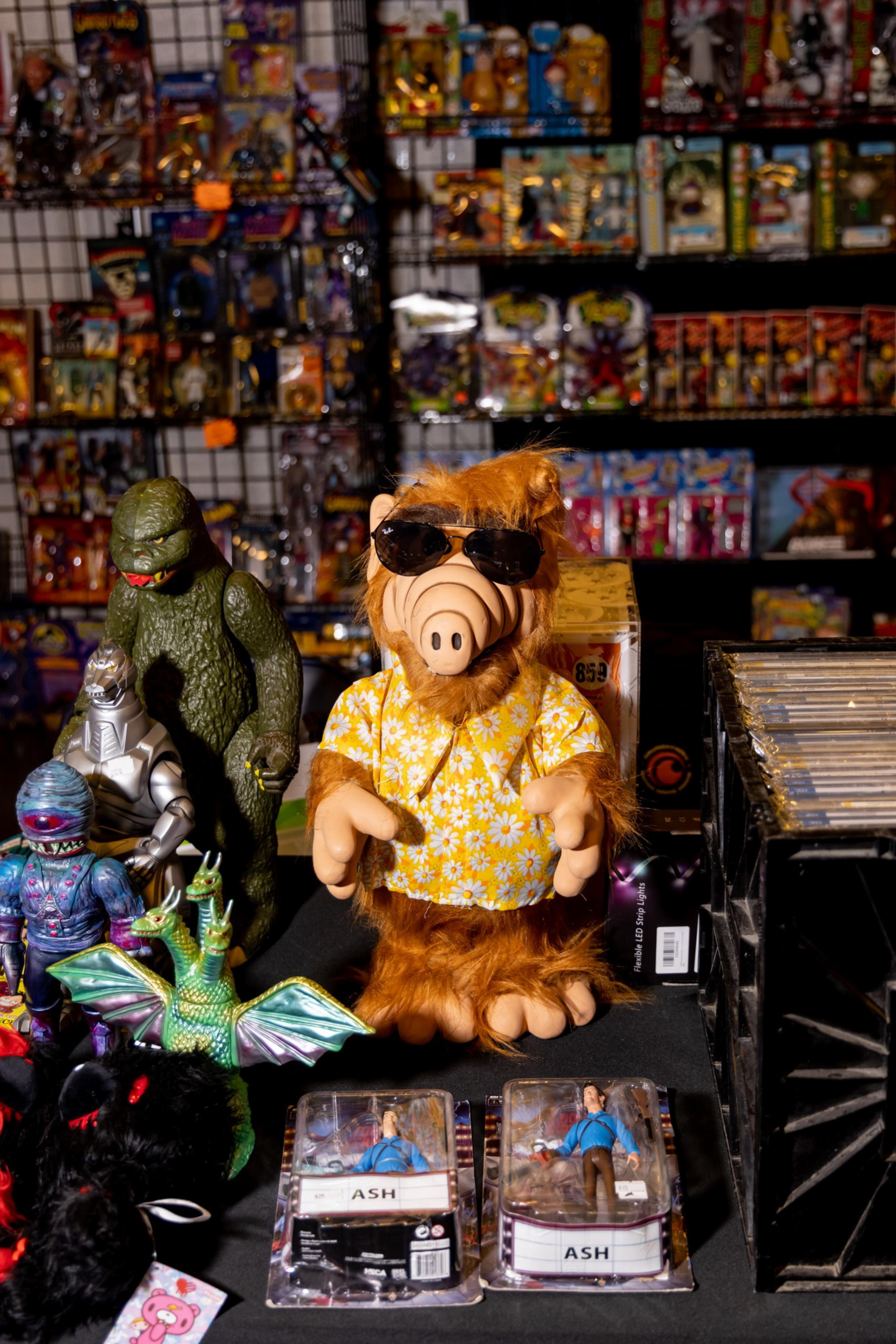A furry character wearing sunglasses and a flower shirt stands among various monster toys and action figures, with shelves of boxed toys in the background.
