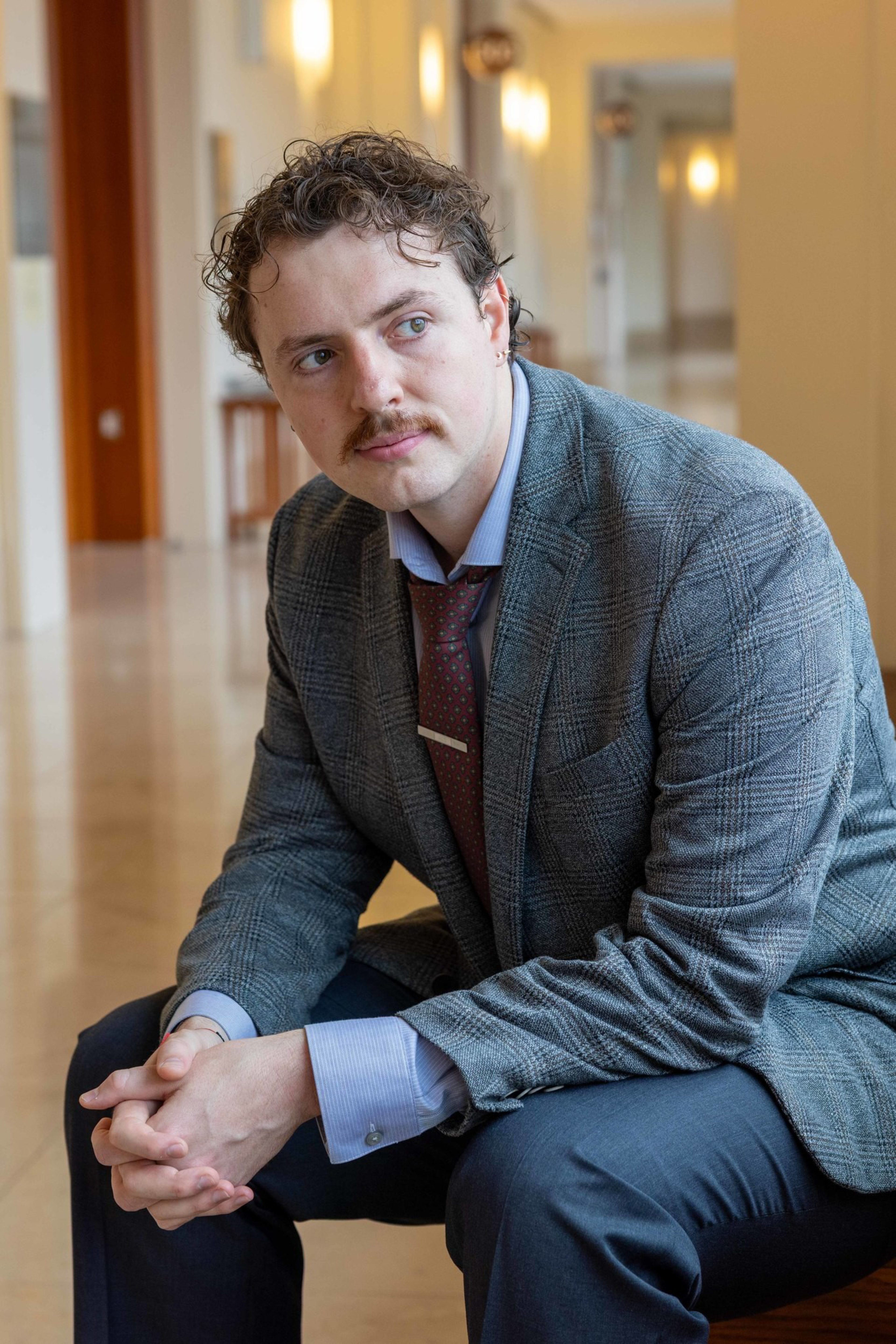 A man with curly hair and a mustache, dressed in a gray checked blazer, blue shirt, and red tie, sits with hands clasped, looking to his left.