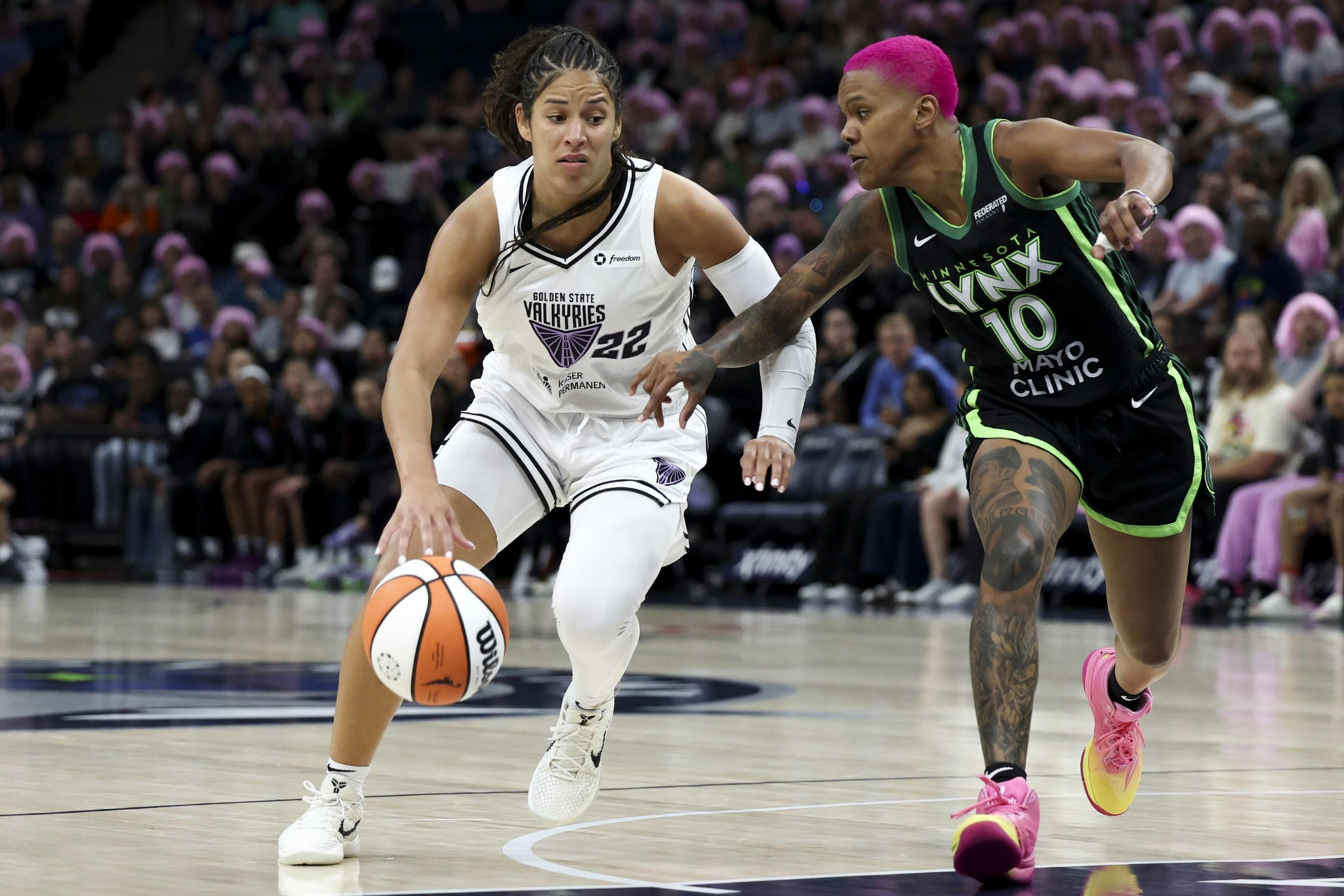A basketball player in a white Golden State Valkyries uniform dribbles the ball while a Minnesota Lynx player with bright pink hair defends closely.