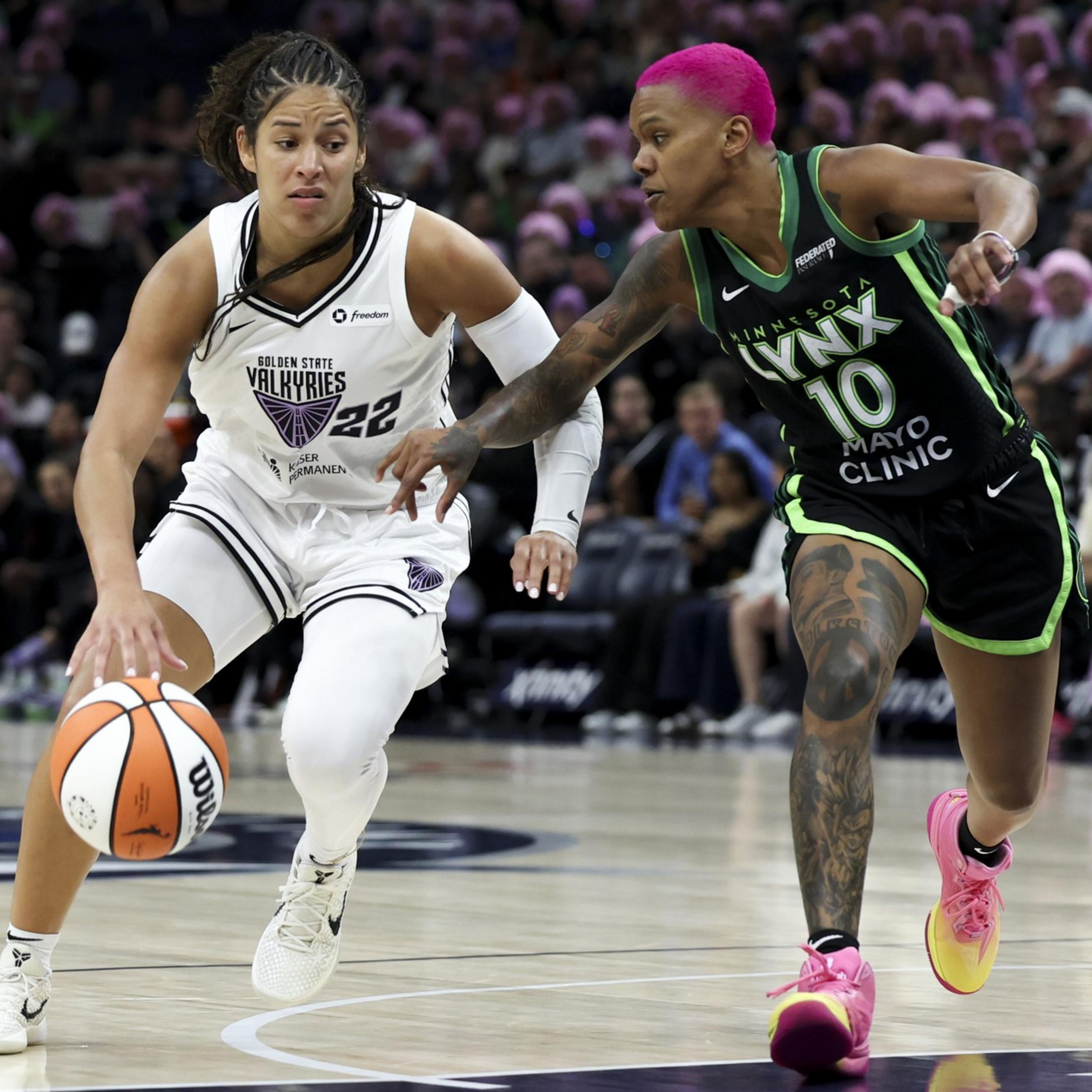 A basketball player in a white Golden State Valkyries uniform dribbles the ball while a Minnesota Lynx player with bright pink hair defends closely.