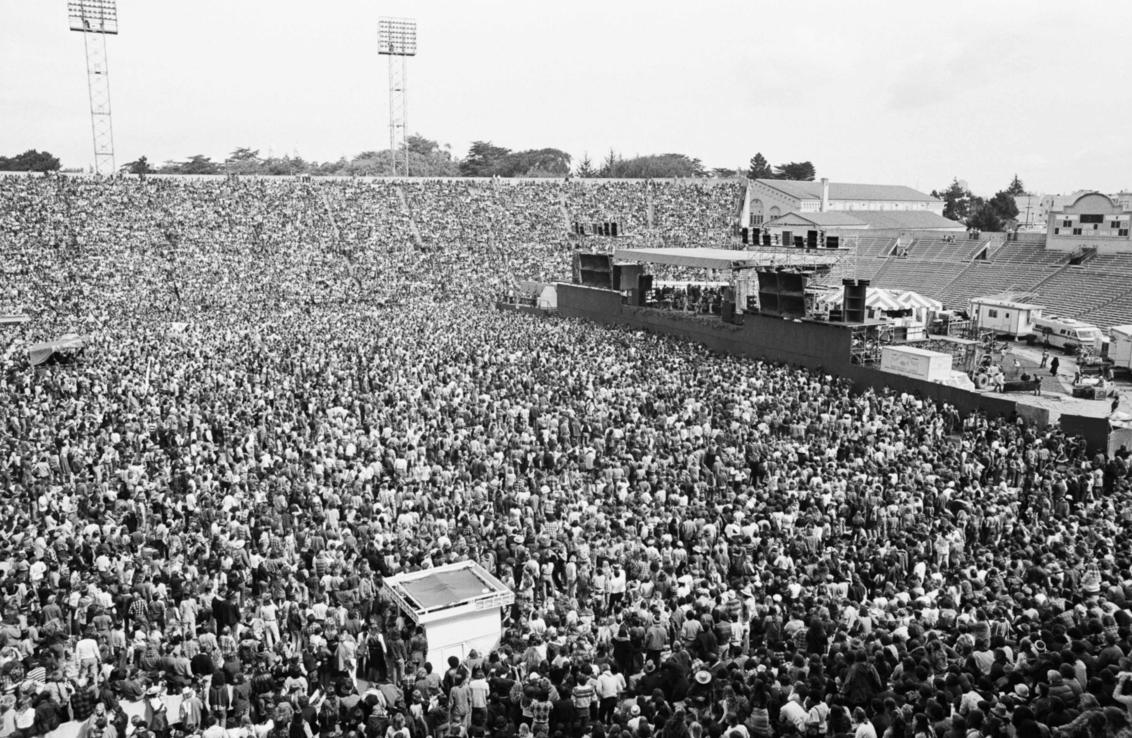 A massive crowd fills an outdoor stadium, densely packed in front of a large stage with speakers and lighting equipment.