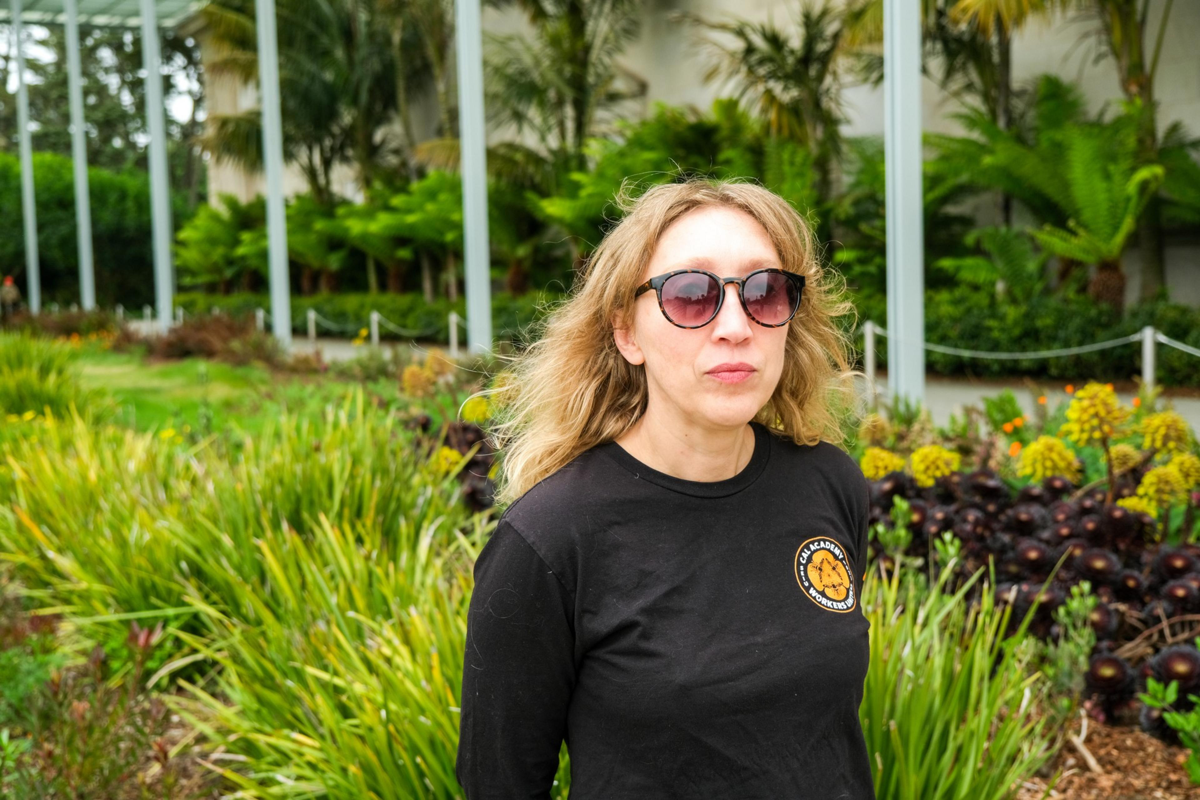 A woman in sunglasses and a black t-shirt stands in front of lush greenery, slightly glancing away from the camera.