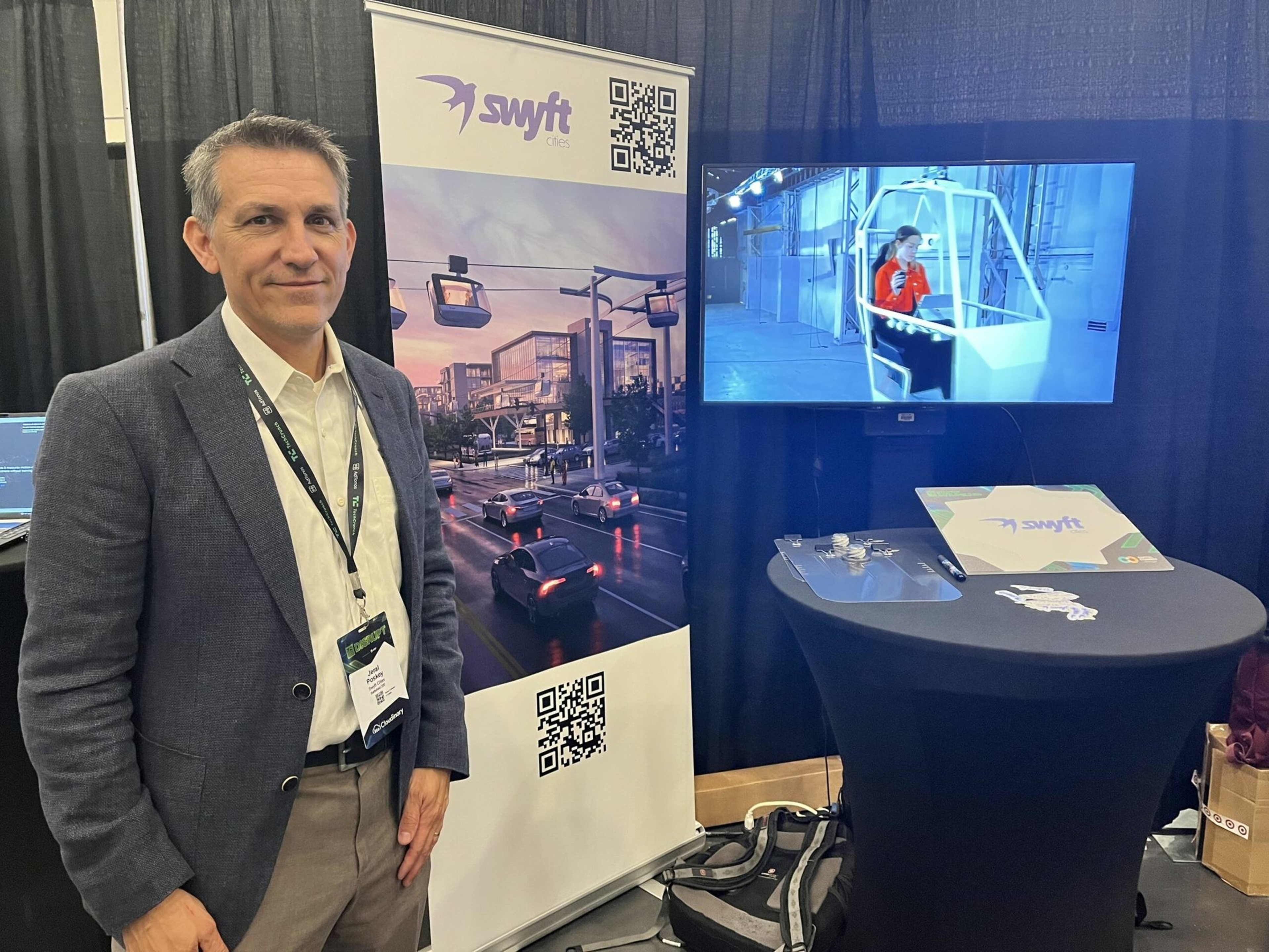 A man in a suit stands next to a banner for "Swyft CES" showing aerial cable cars over a city street, with a monitor and table displaying related items nearby.