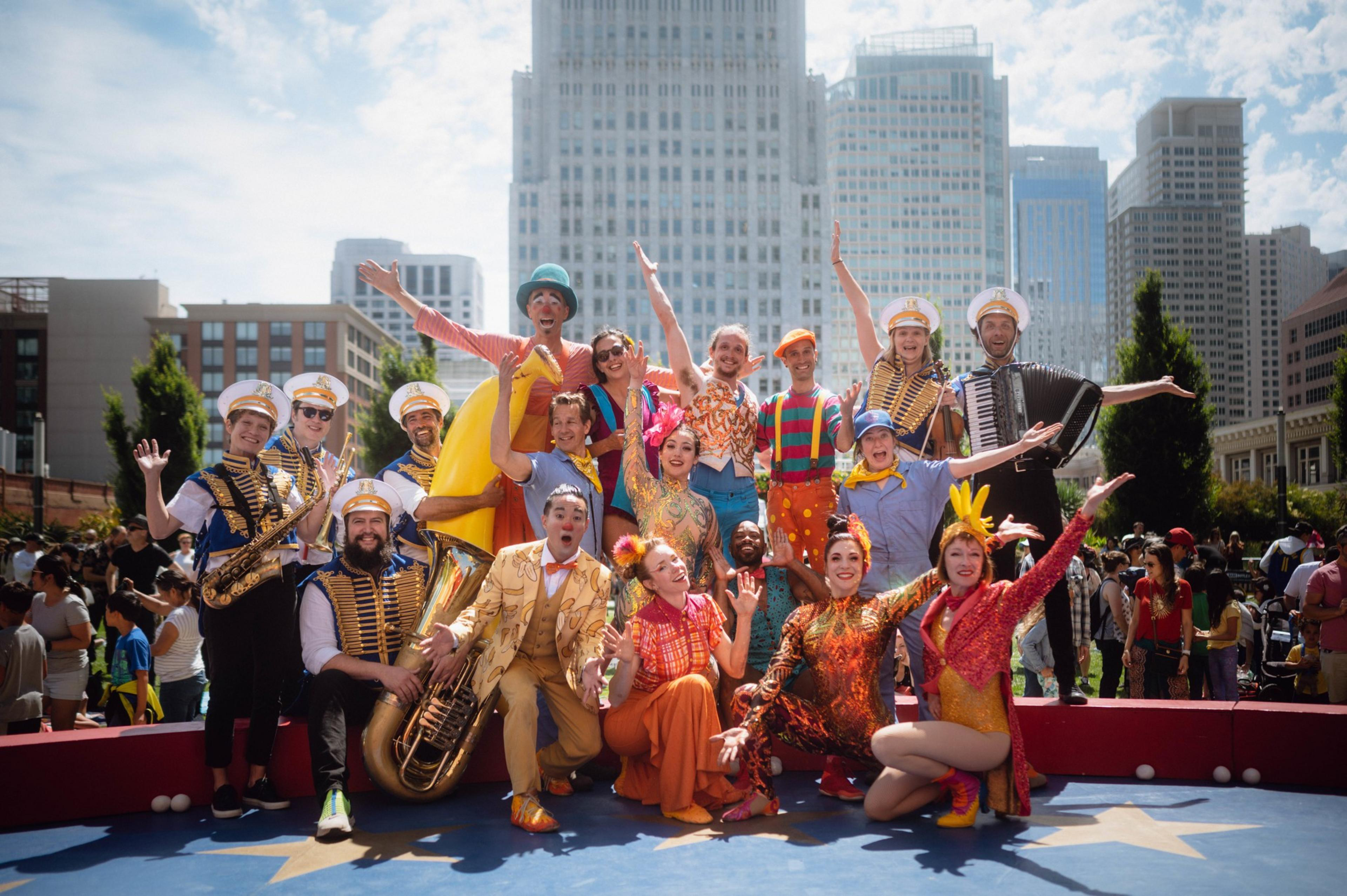 A lively group of performers in colorful costumes and hats pose energetically in front of a cityscape, with some playing musical instruments.