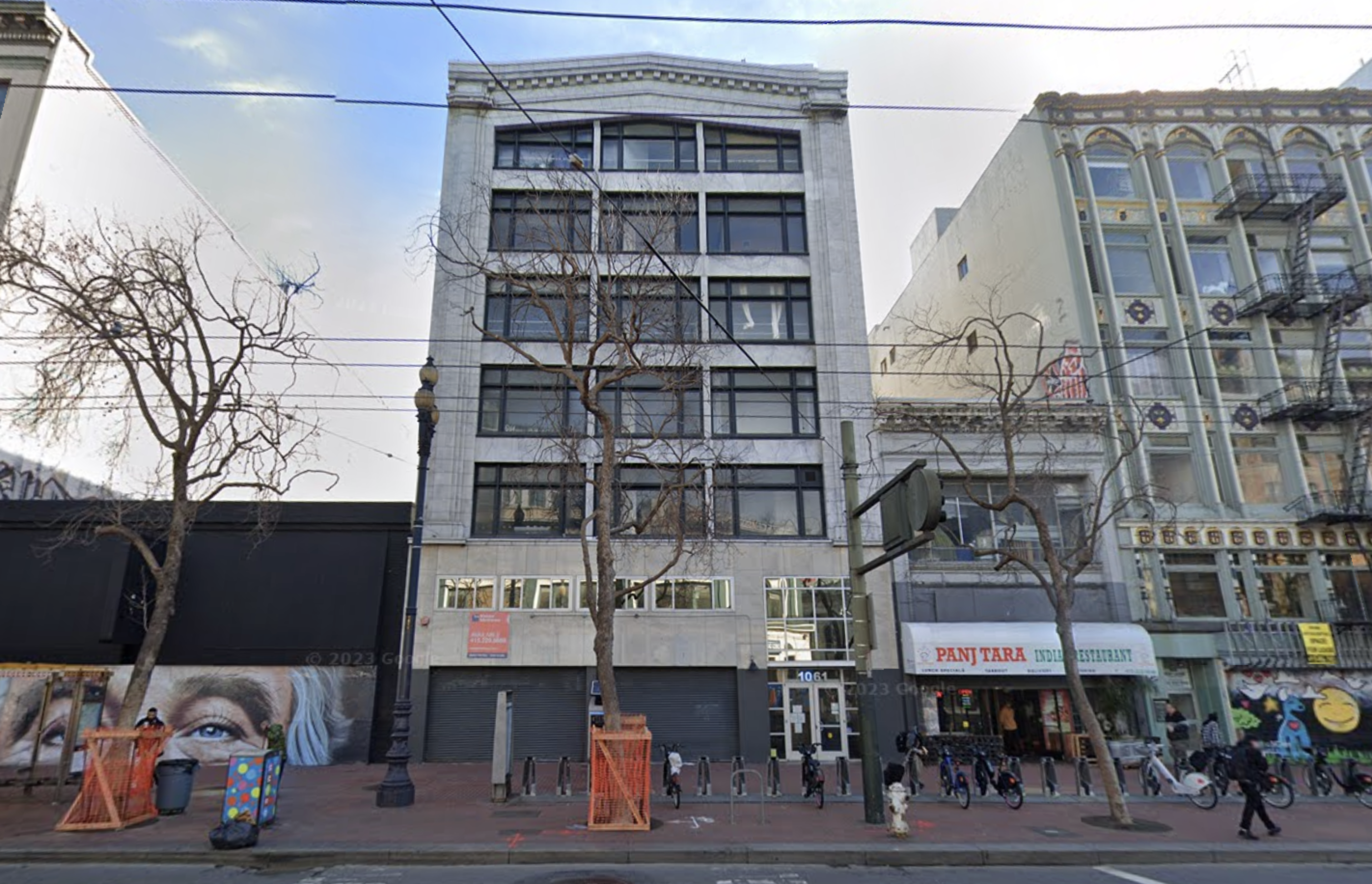 A six-story building with a gray facade stands between two older buildings, one with a mural and the other hosting a restaurant named Panj Tara.