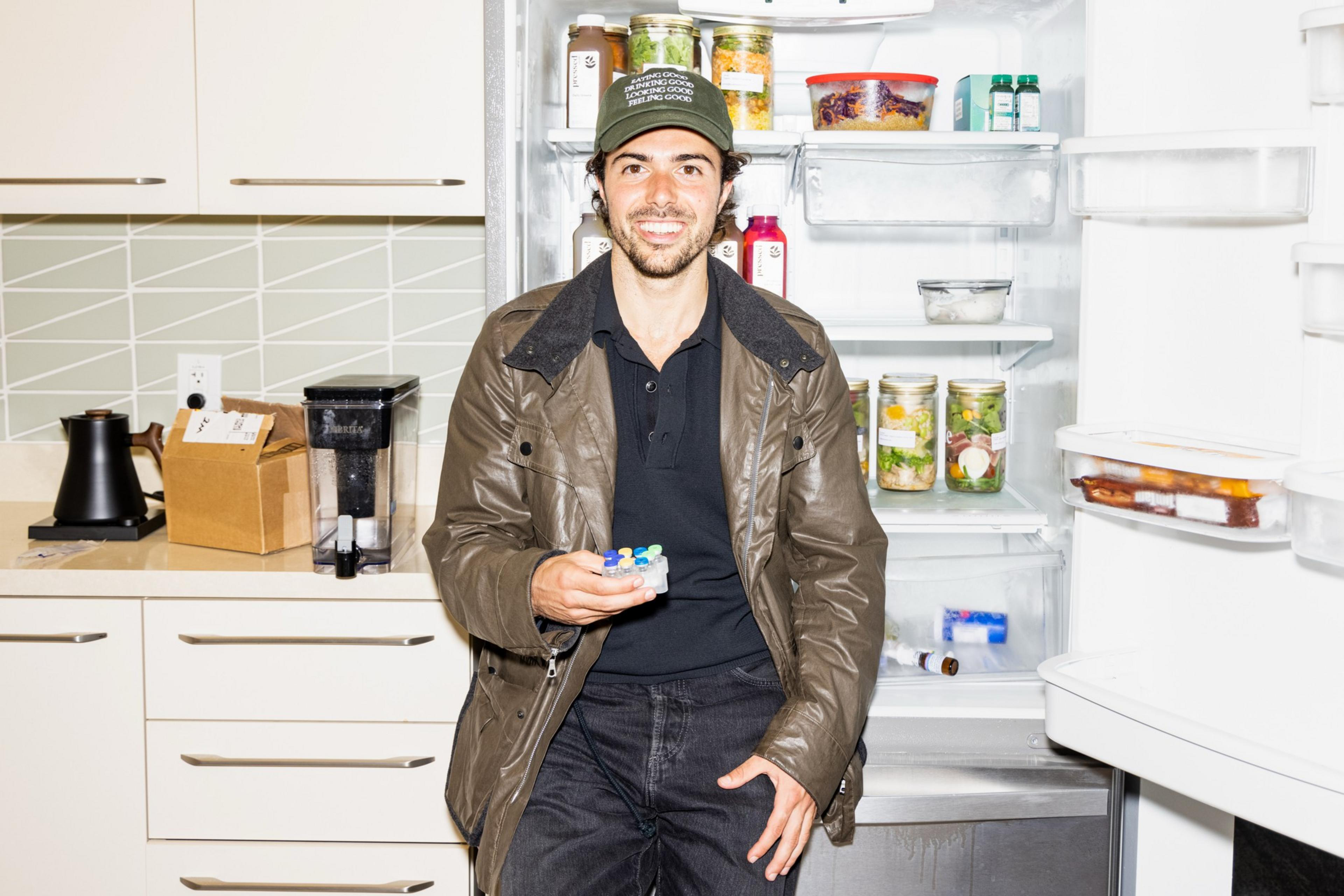 A man wearing a brown jacket and green hat sits by an open fridge holding a small container with colorful vape pens inside.