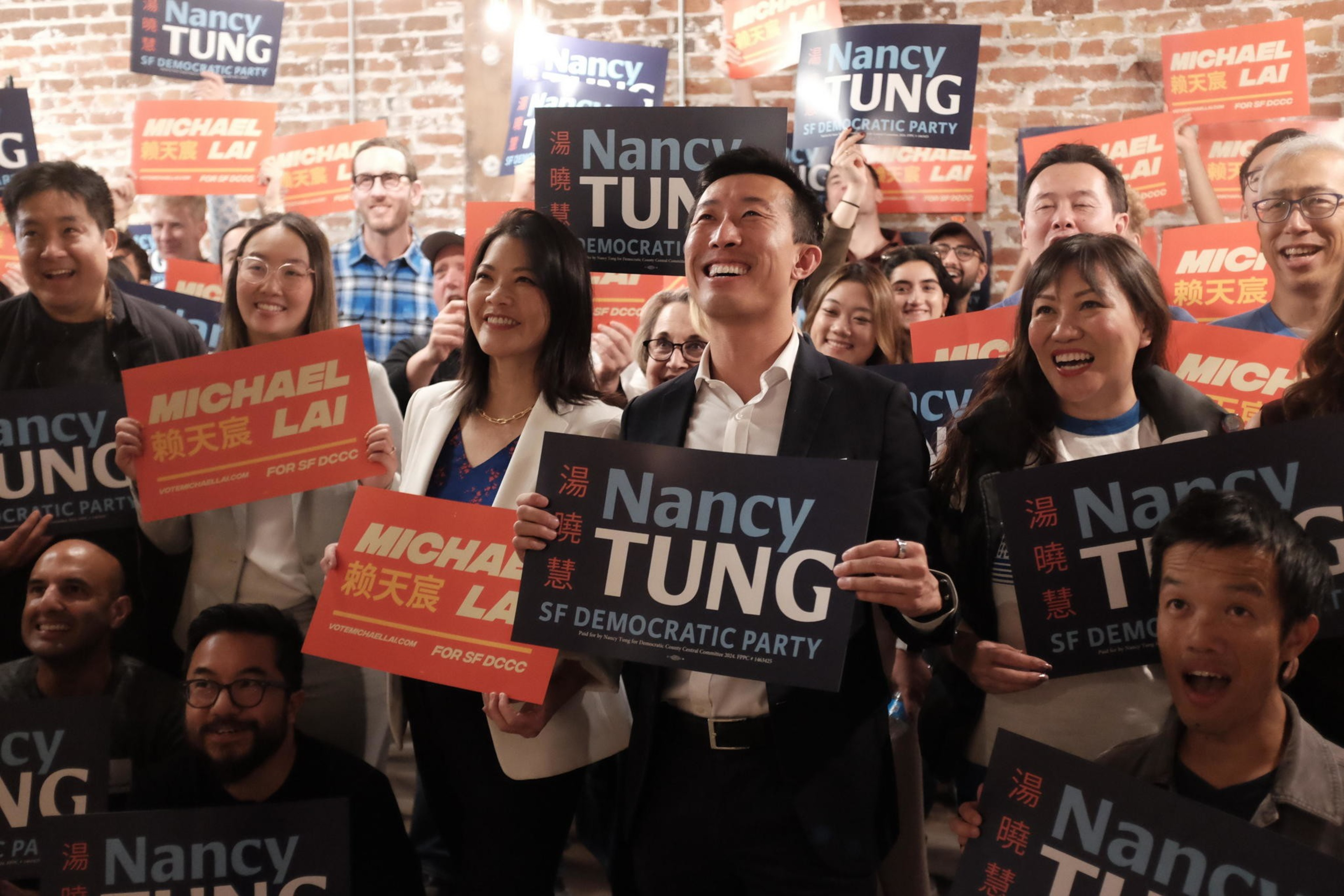 A group of moderate Democrats, holding signs, smile against a brick wall at a launch party for a moderate slate.