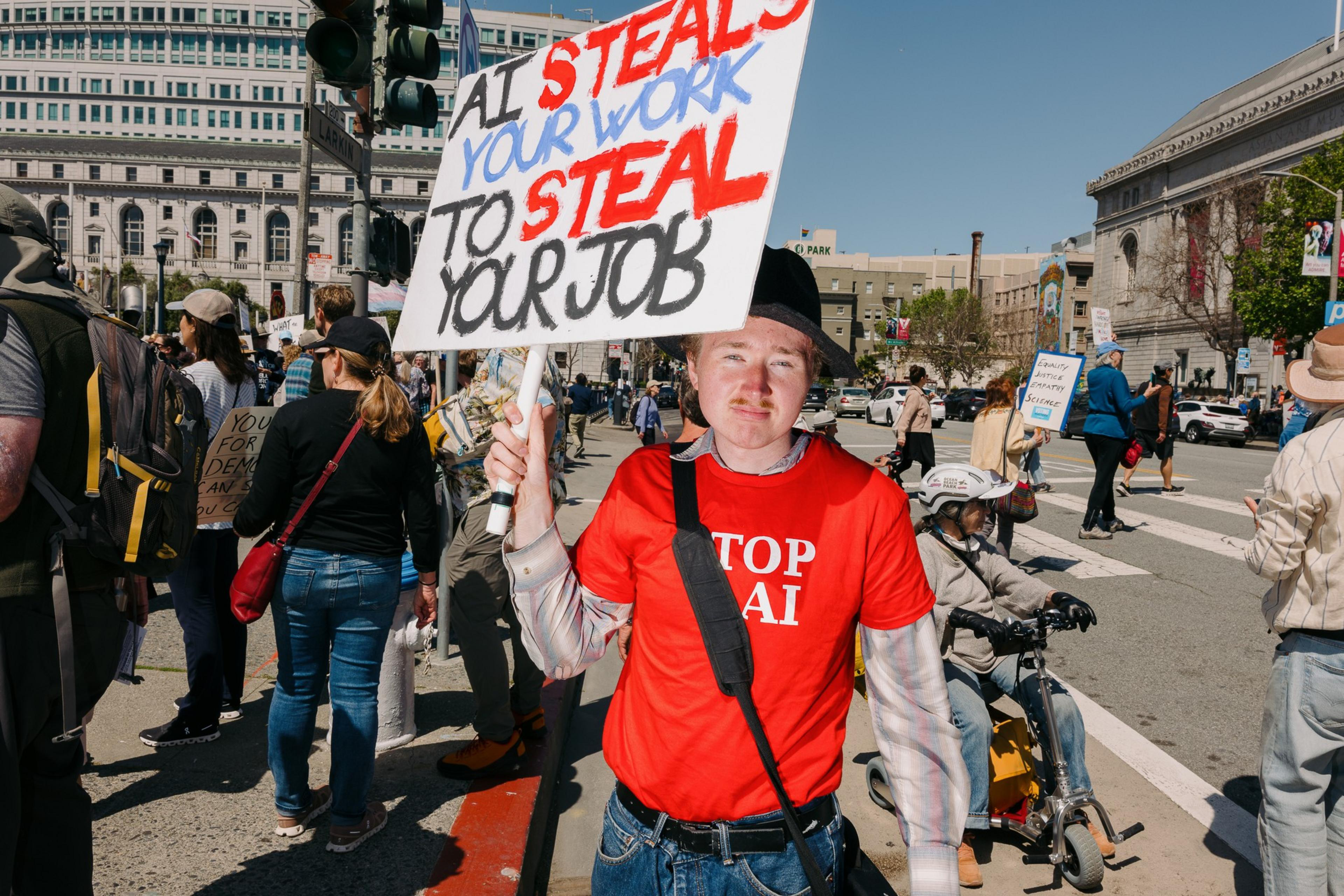 A person in a red &quot;Stop AI&quot; shirt holds a sign reading &quot;AI steals your work to steal your job,&quot; standing amidst a crowd at a protest on a city street.