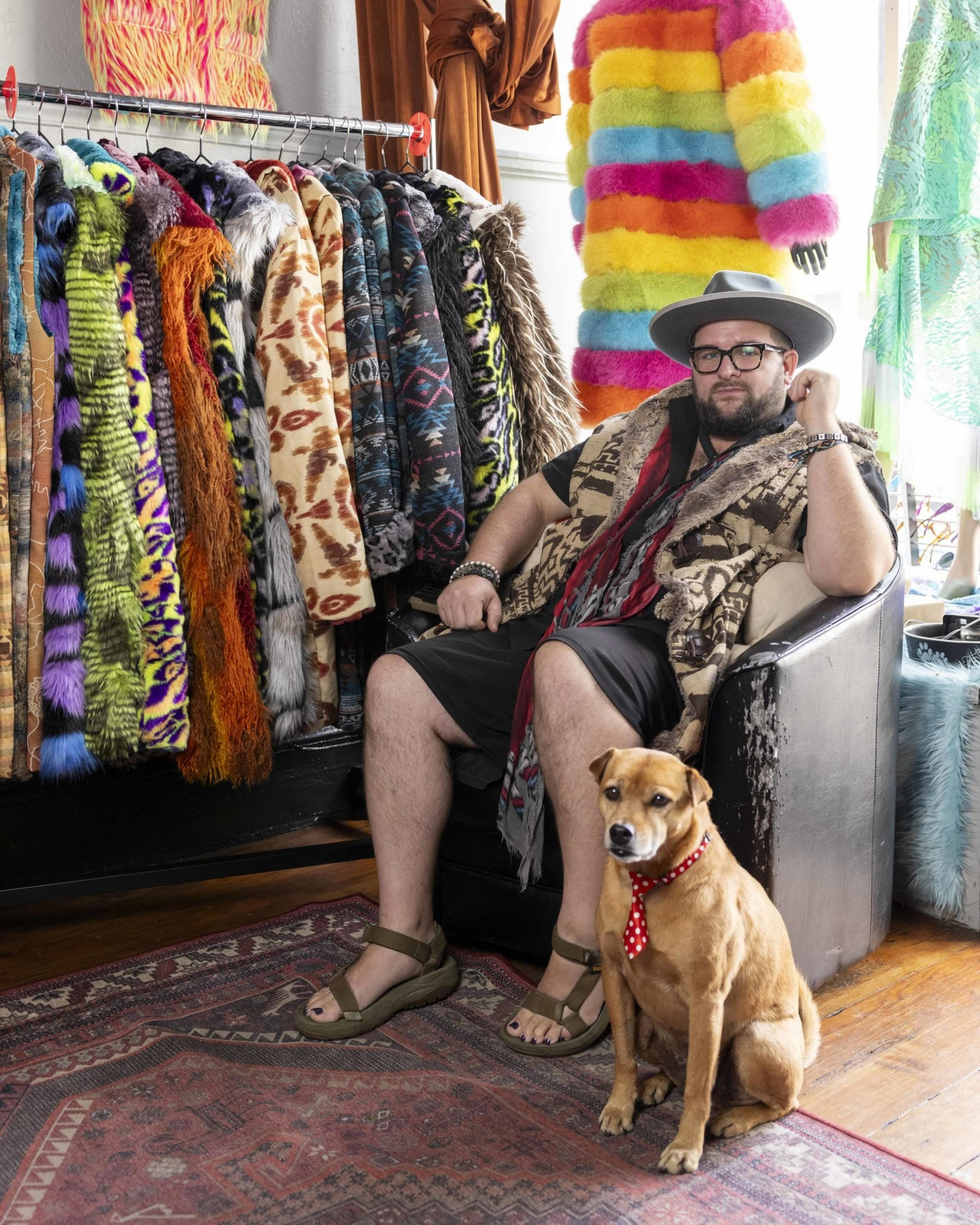 A bearded man in a hat and sandals sits in a chair next to a dog with a red polka dot bandana, surrounded by colorful fur coats and outfits on racks and mannequins.