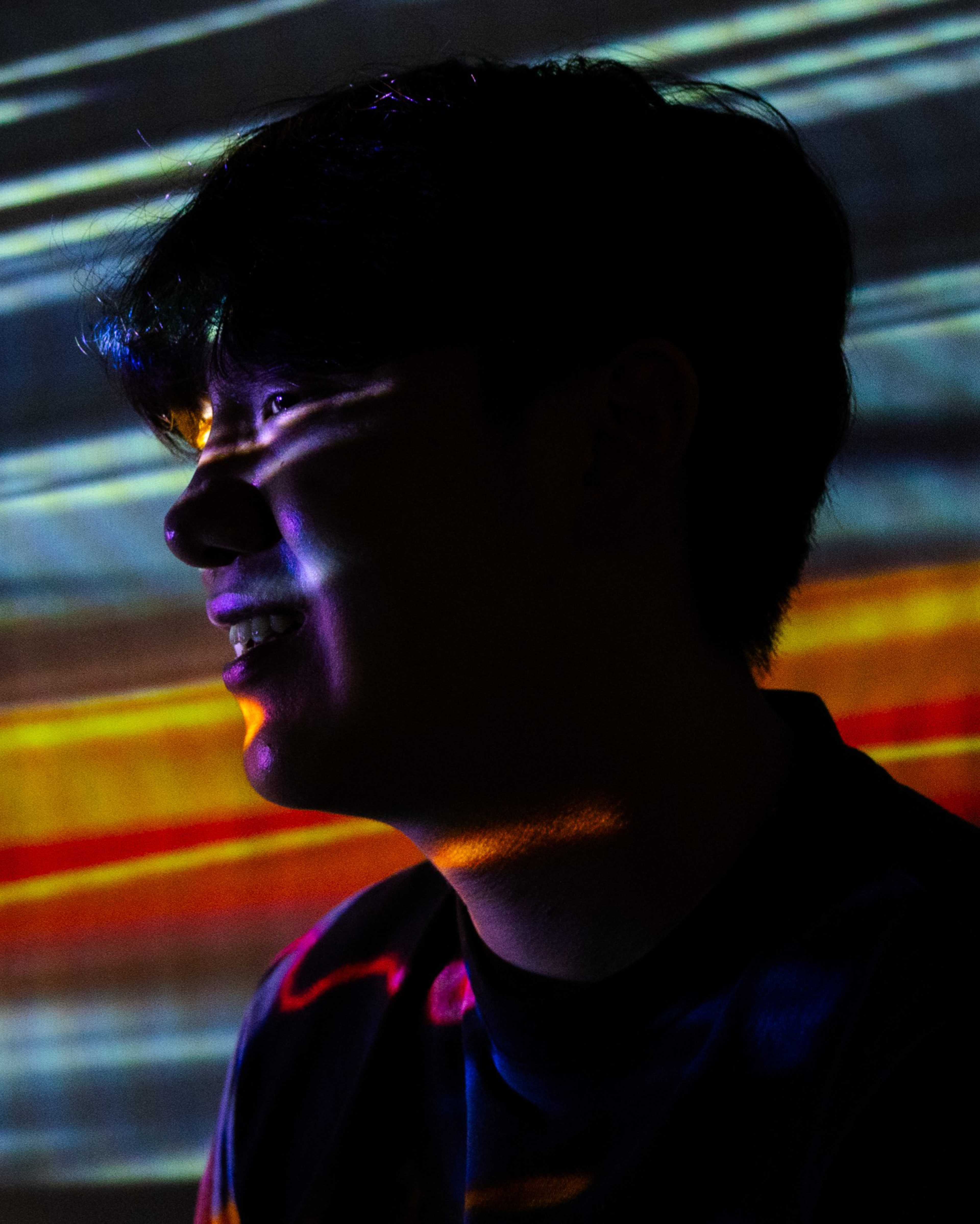 A person’s side profile is lit by multicolored light patterns, casting blue, purple, yellow, and red hues across their face and background.