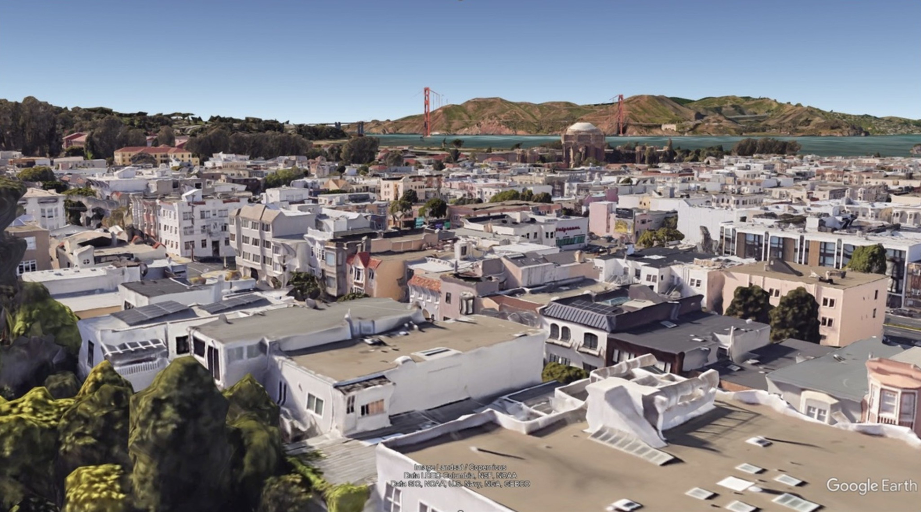 This image shows an aerial view of a densely packed city neighborhood with houses and buildings, trees, and a bridge in the background stretching over a body of water.
