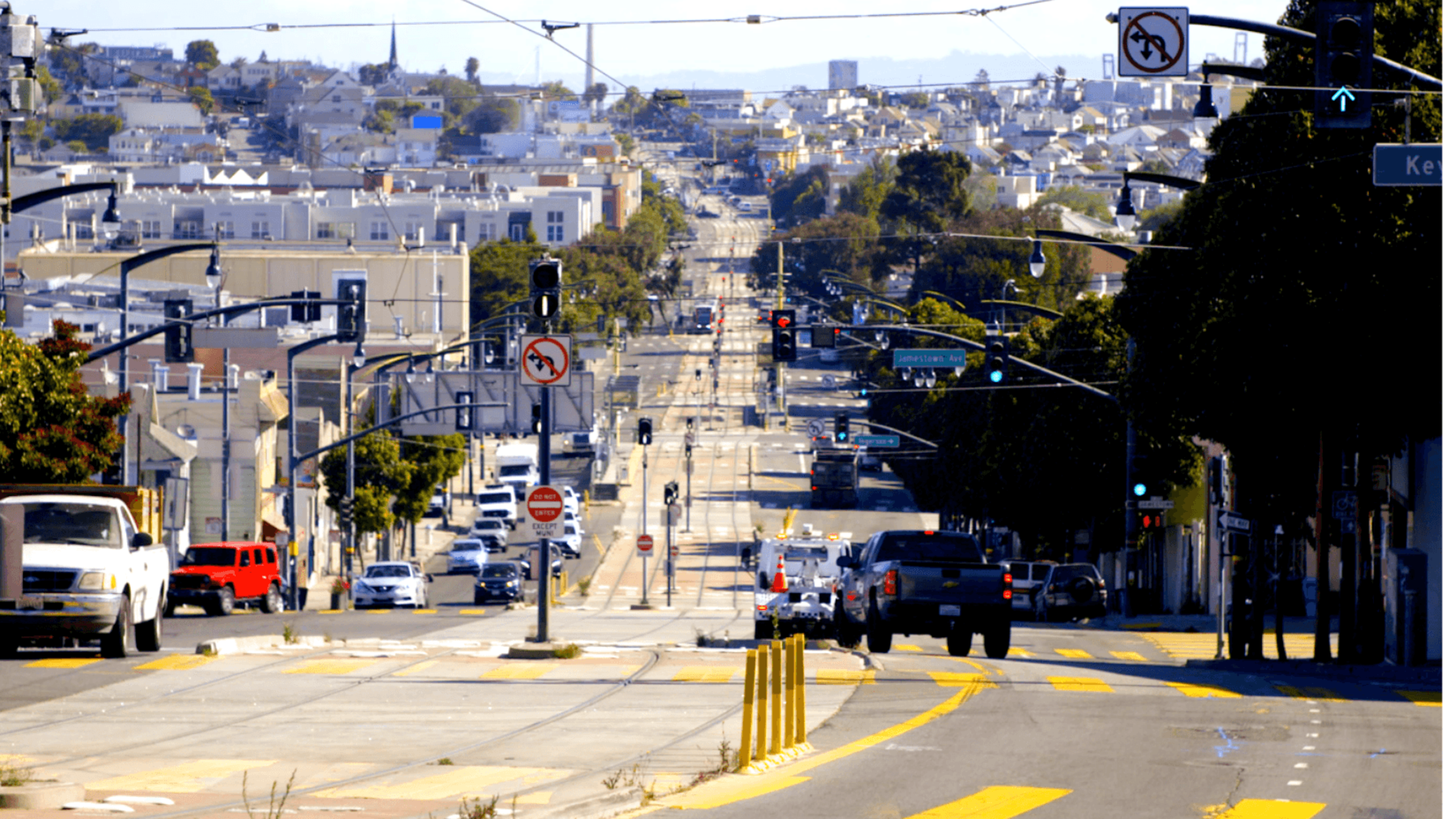 A city street with cars driving along, traffic lights, and no-left-turn signs, stretching into the distance with buildings and trees on both sides.