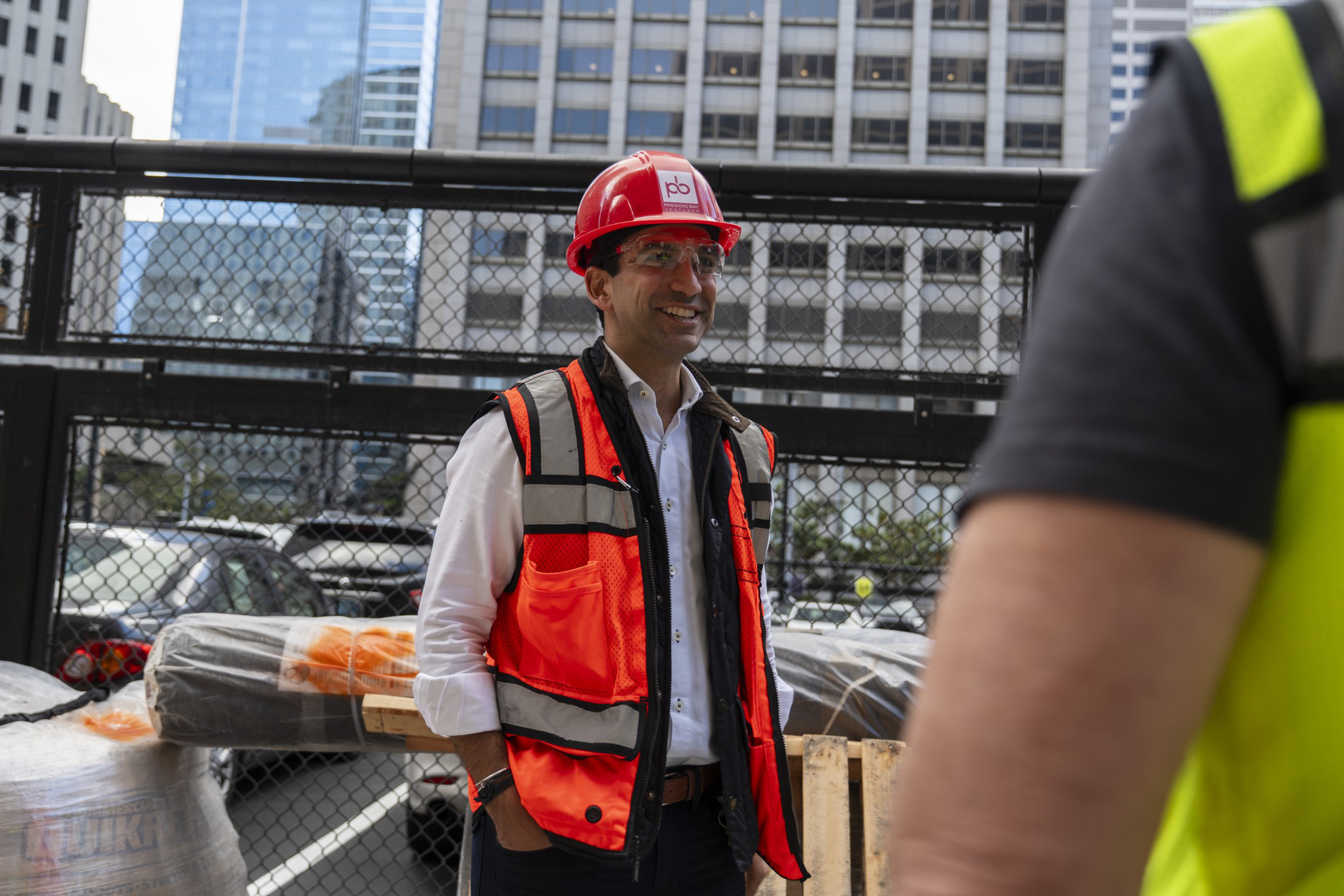 A man in a red hard hat and orange safety vest smiles while standing near construction materials, with city buildings and a fenced area behind him.