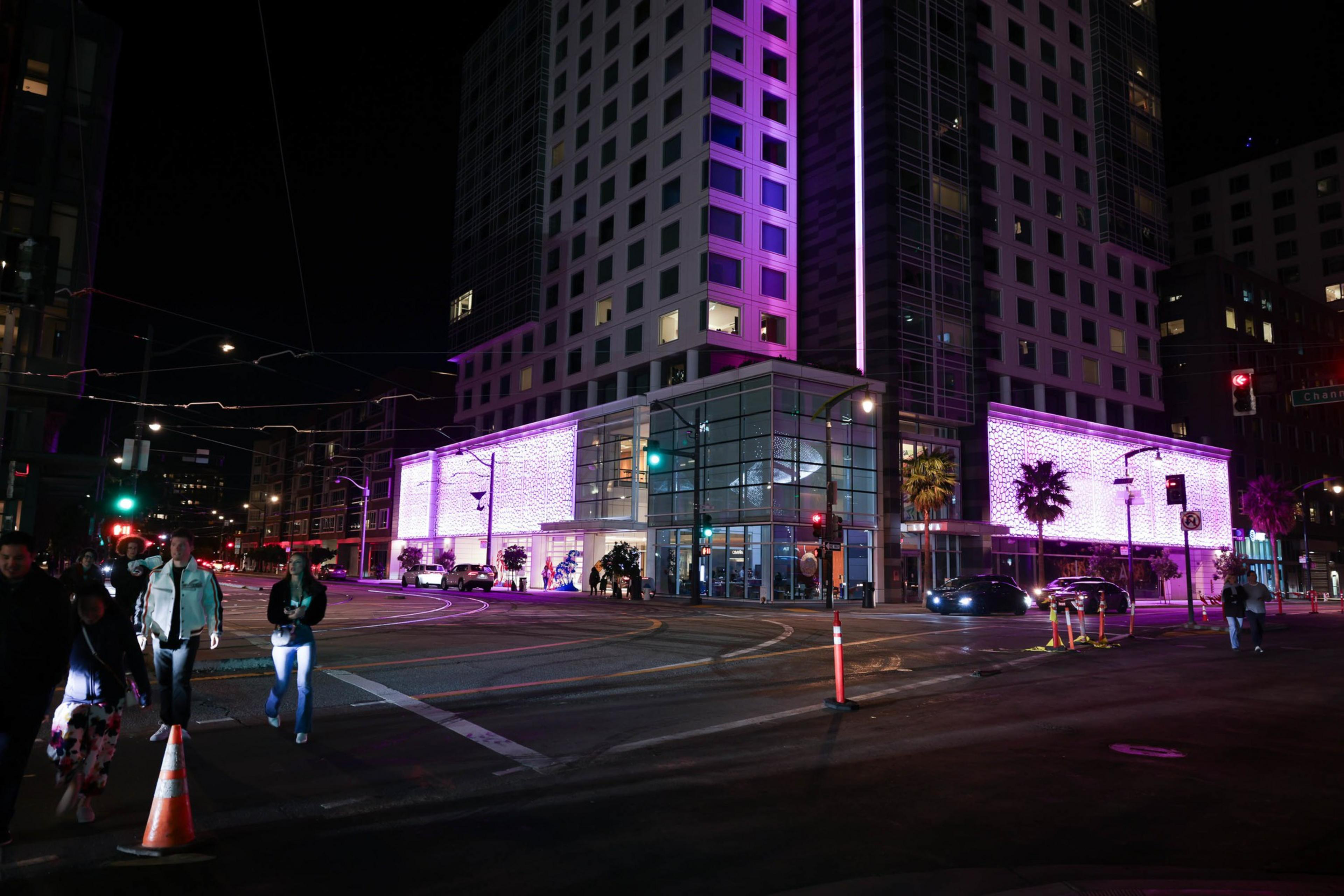 A city street at night with pedestrians crossing, illuminated buildings, and palm trees.