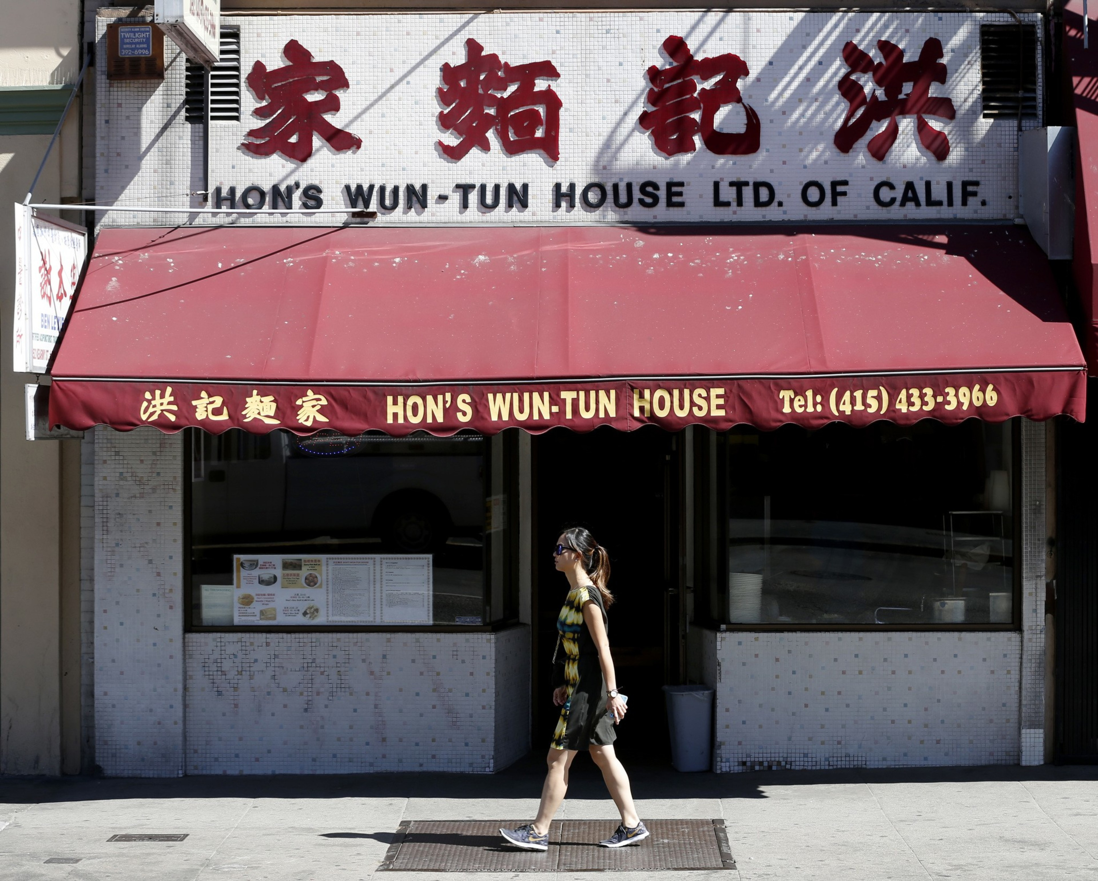 A woman in a patterned dress and sneakers walks past Hon's Wun-Tun House, a restaurant with a red awning and Chinese characters on its sign.