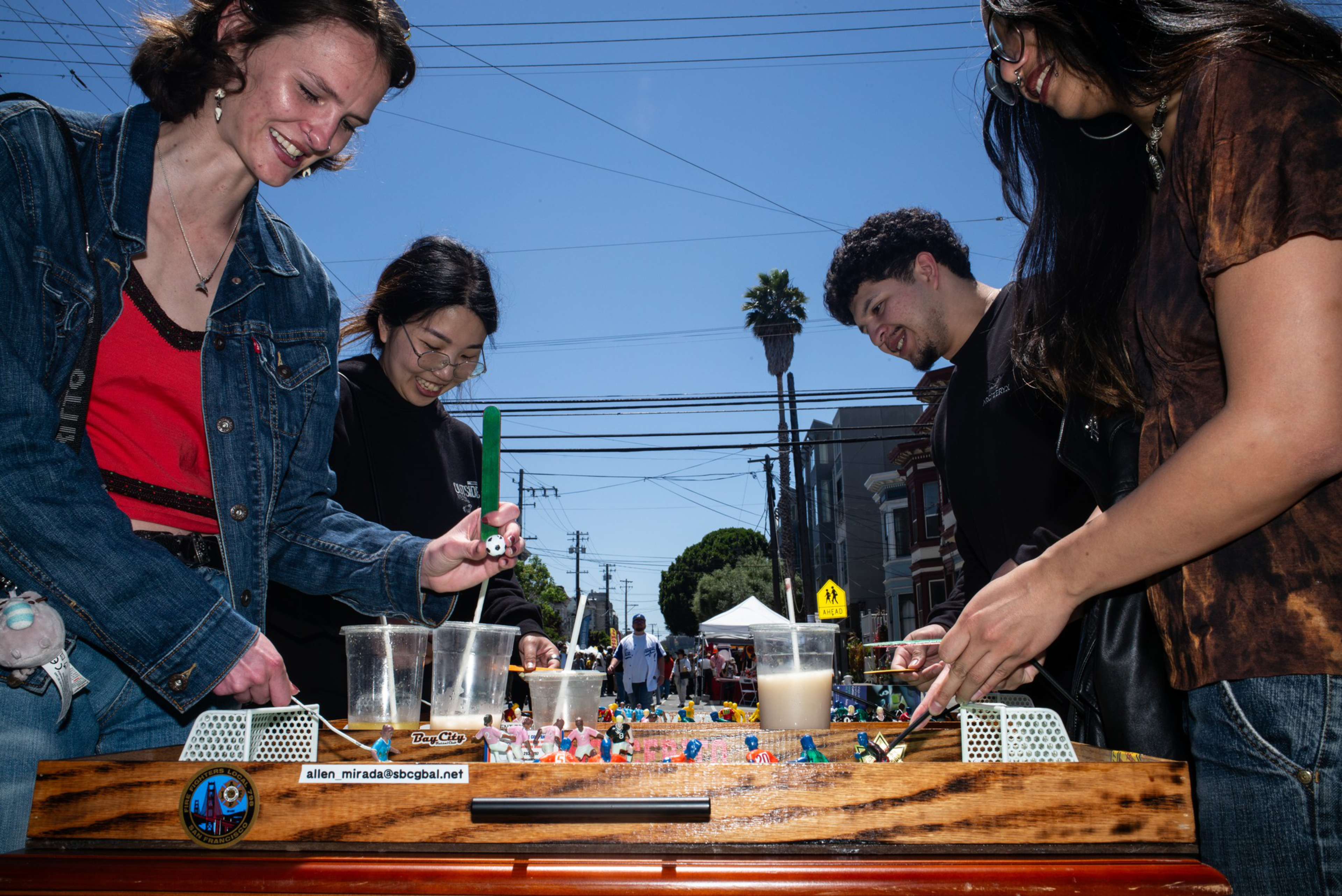 Four people stand around a foosball table outside, smiling and playing, with drinks placed on the table under a clear blue sky.