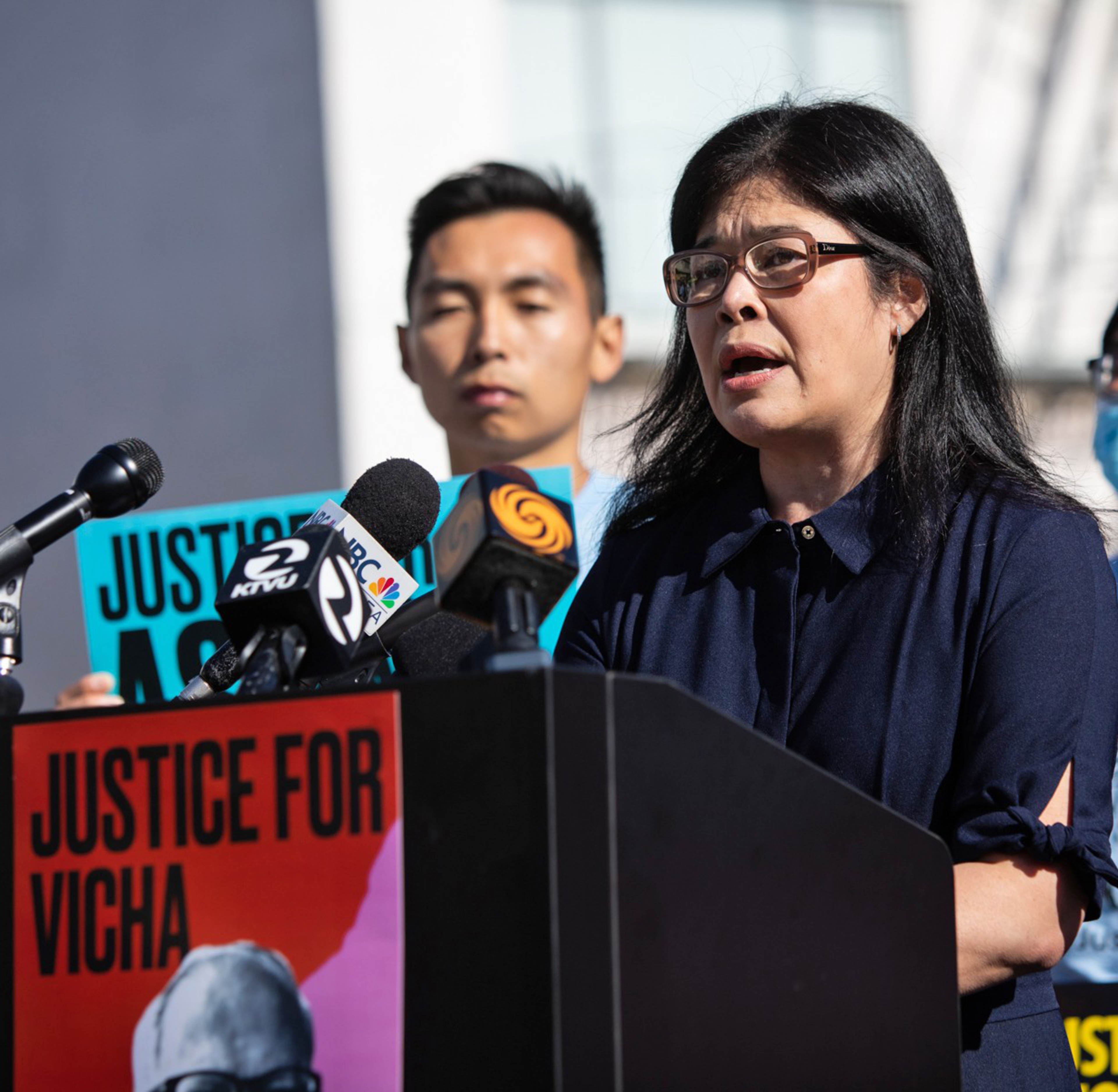A woman stands at a podium speaking into microphones labeled with news logos, while a man behind her holds a blue sign reading "JUSTICE."