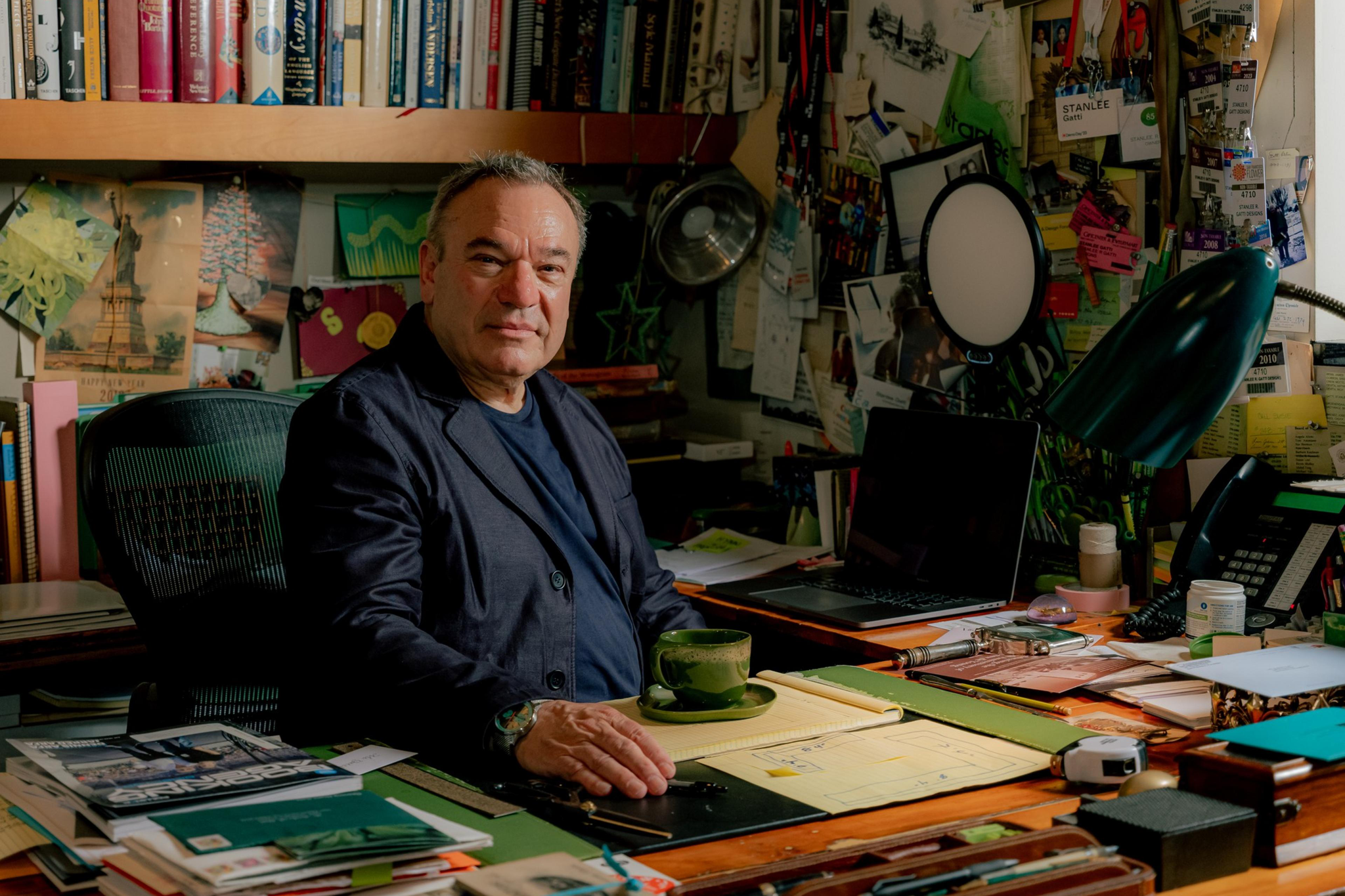 A man in a dark jacket sits at a cluttered desk filled with papers, books, and a green mug, surrounded by shelves and walls covered with photos, notes, and badges.