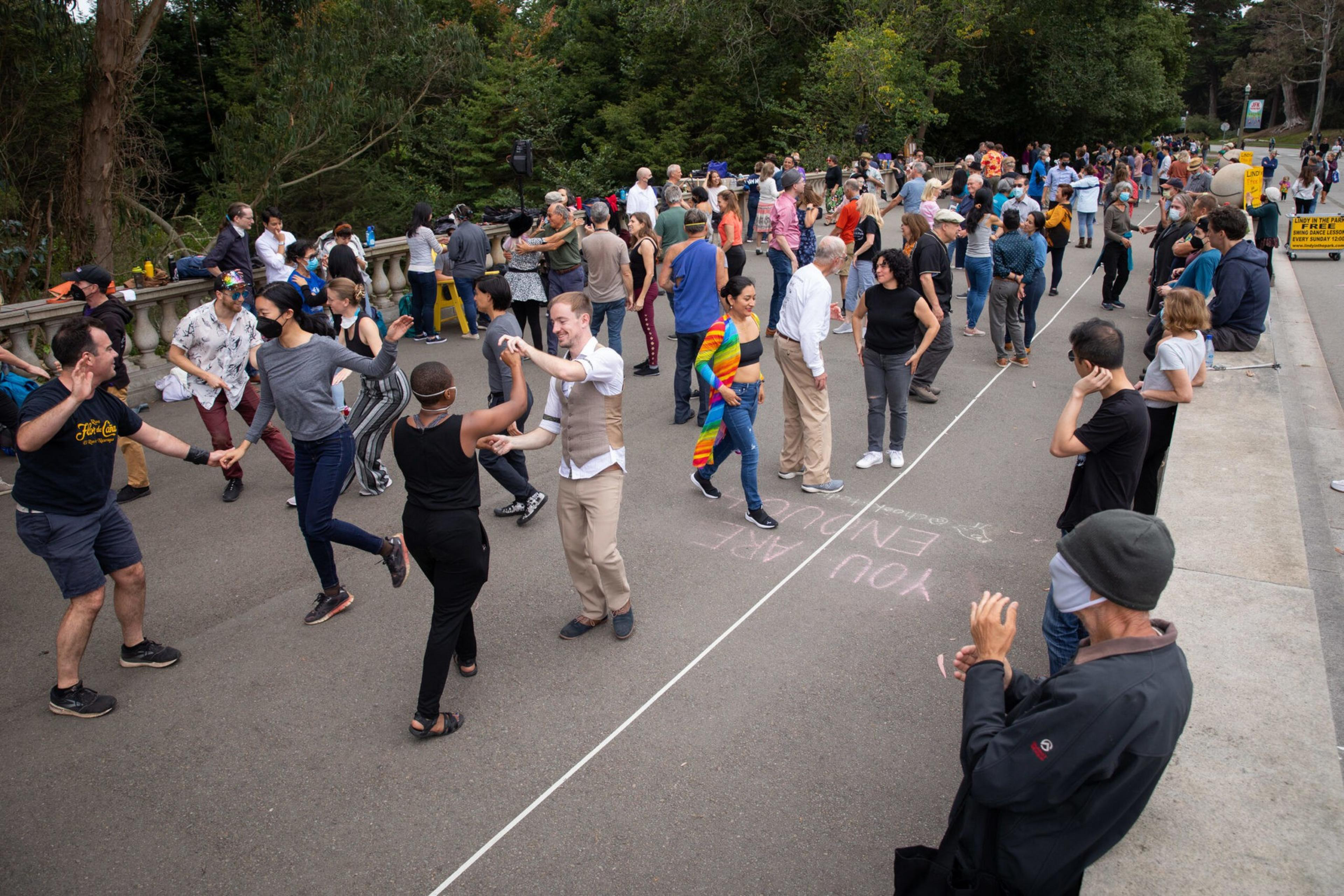 A diverse group of people dance together outside on a road, with some lined up along a chalk line marked "YOU ARE HERE."