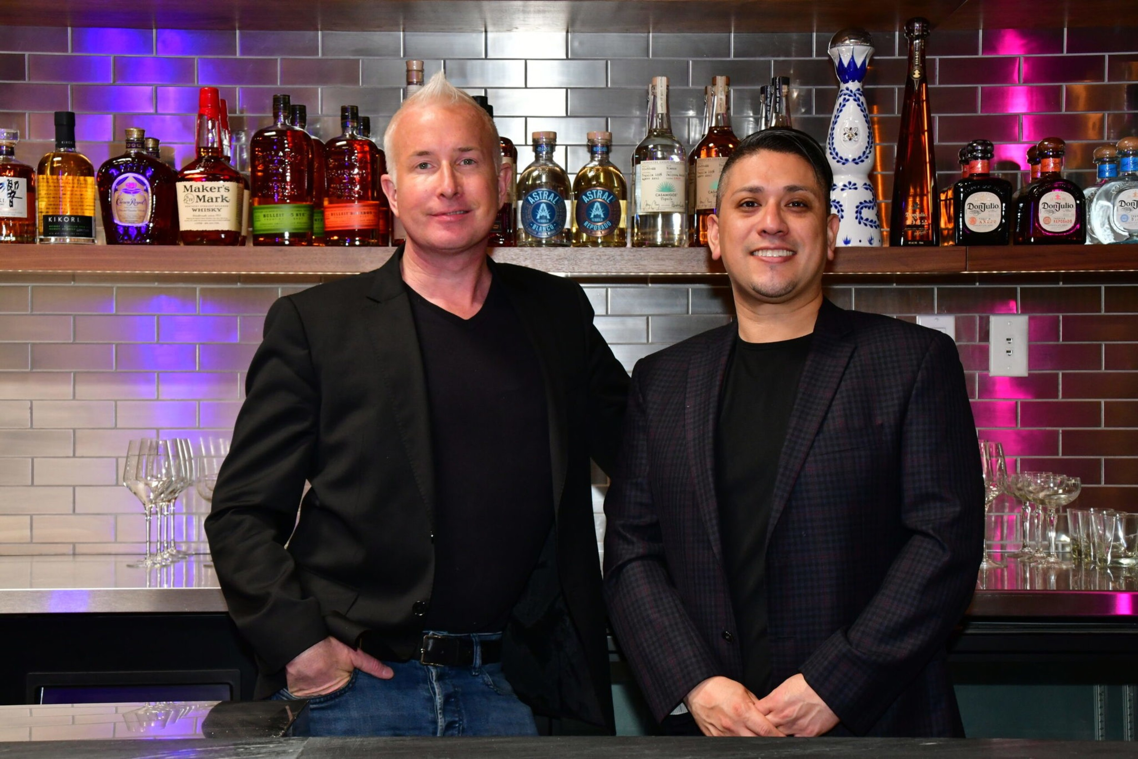 Two men in dark blazers stand behind a bar counter with shelves of liquor bottles and glassware on a tiled wall with purple lighting.