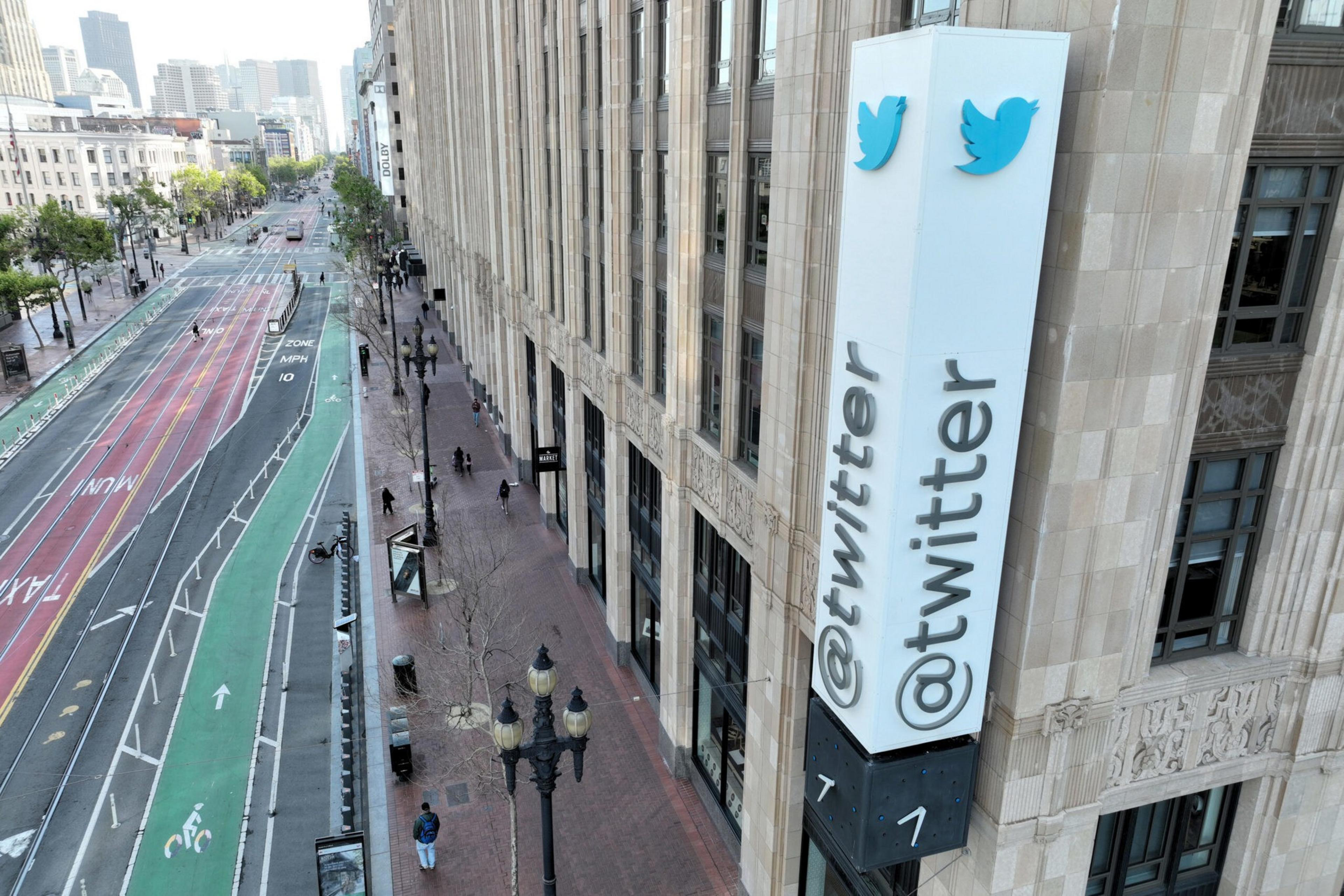 A large Twitter sign with the handle "@twitter" and the blue bird logo is mounted on a tall building next to a city street with bike lanes.