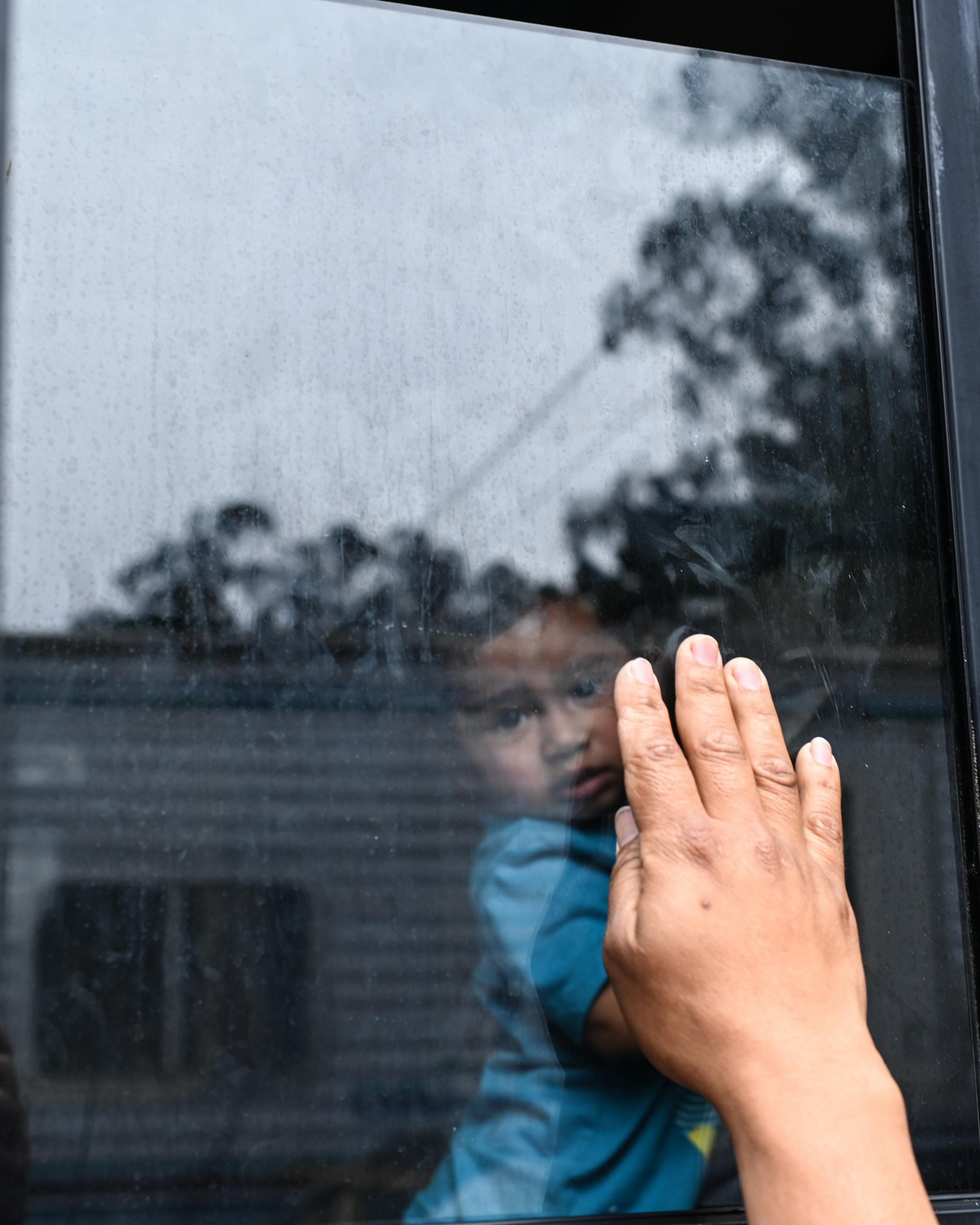 A hand touches a glass window, and through the glass, the reflection of a young child in a blue shirt can be seen, looking towards the camera.