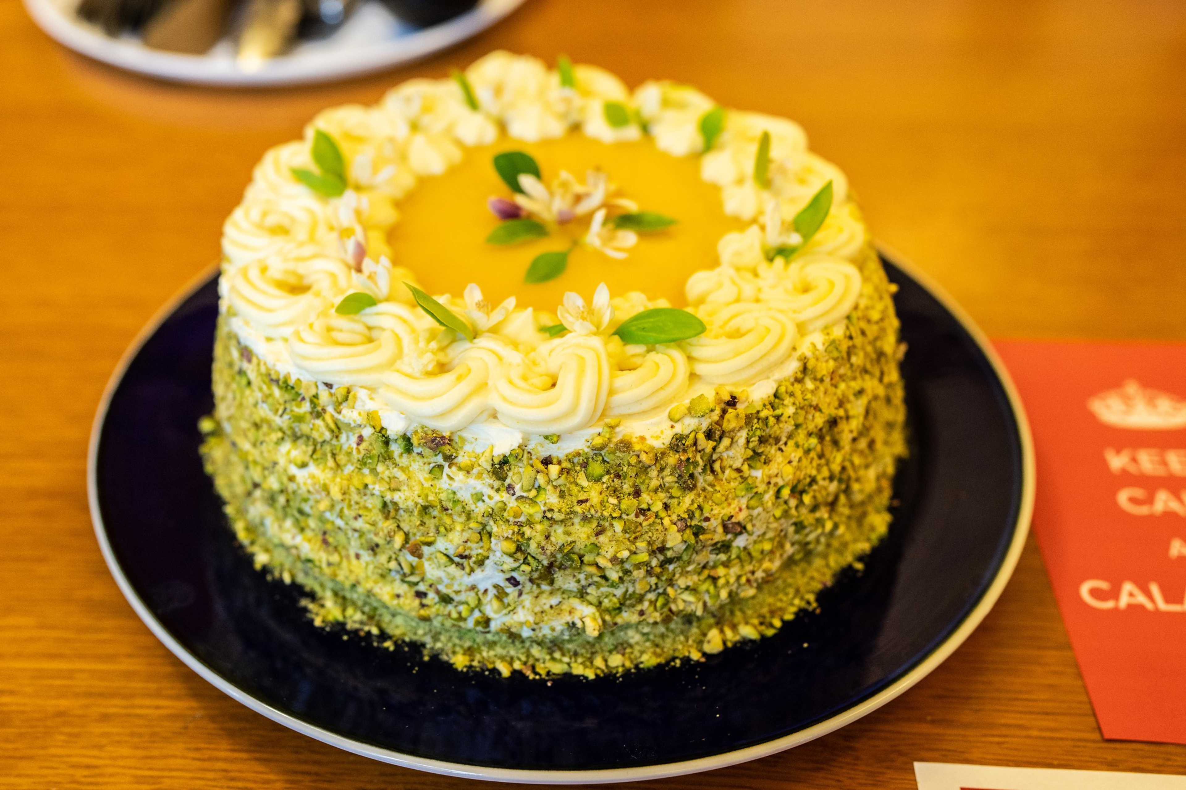 A round cake with white frosting, green nut crumbs on the sides, a yellow glaze center, and small green leaves and flowers as decoration on top sits on a dark plate.
