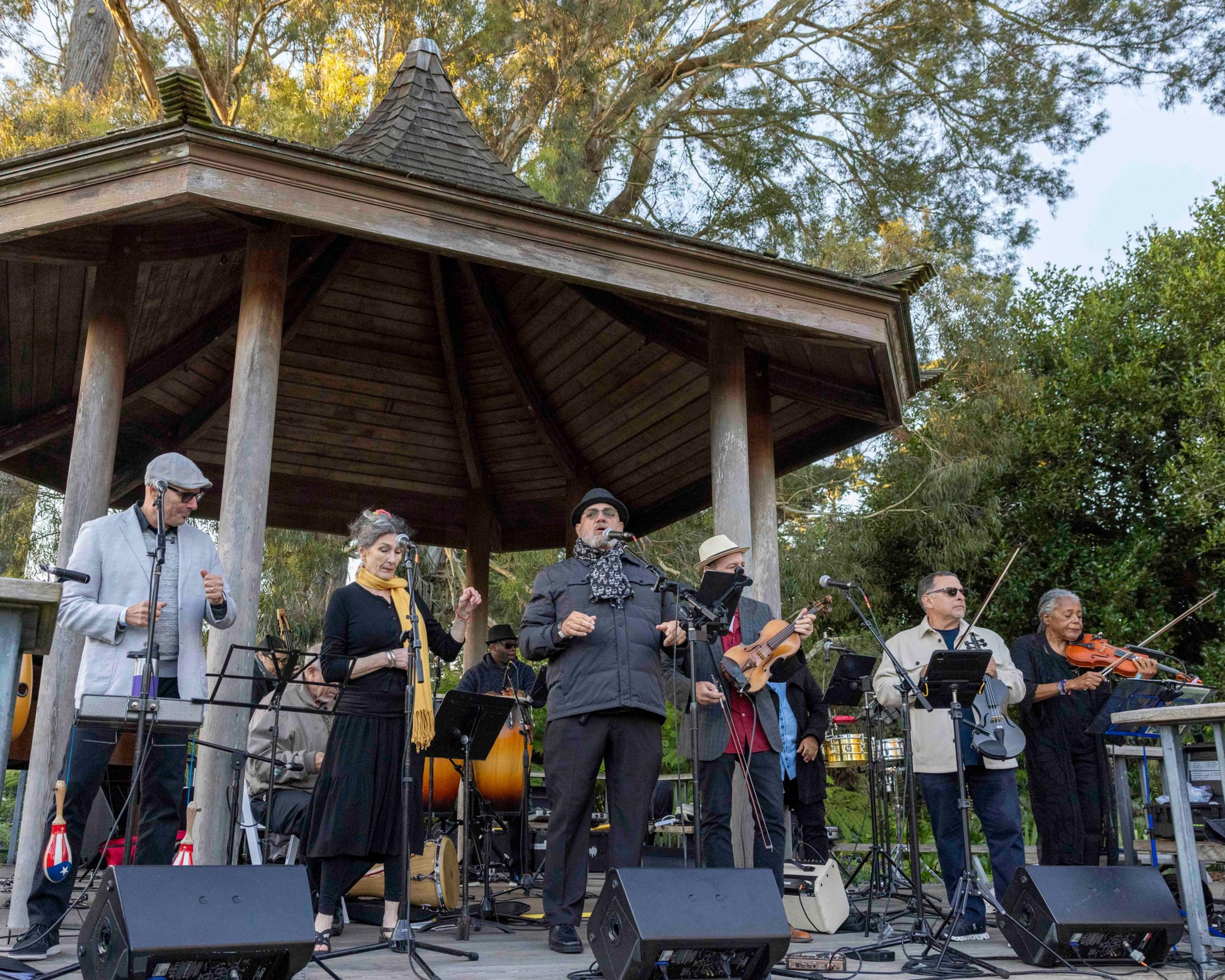 A diverse group of musicians performs under a wooden gazebo, surrounded by trees. They play various instruments and sing, dressed in casual attire with hats and scarves.