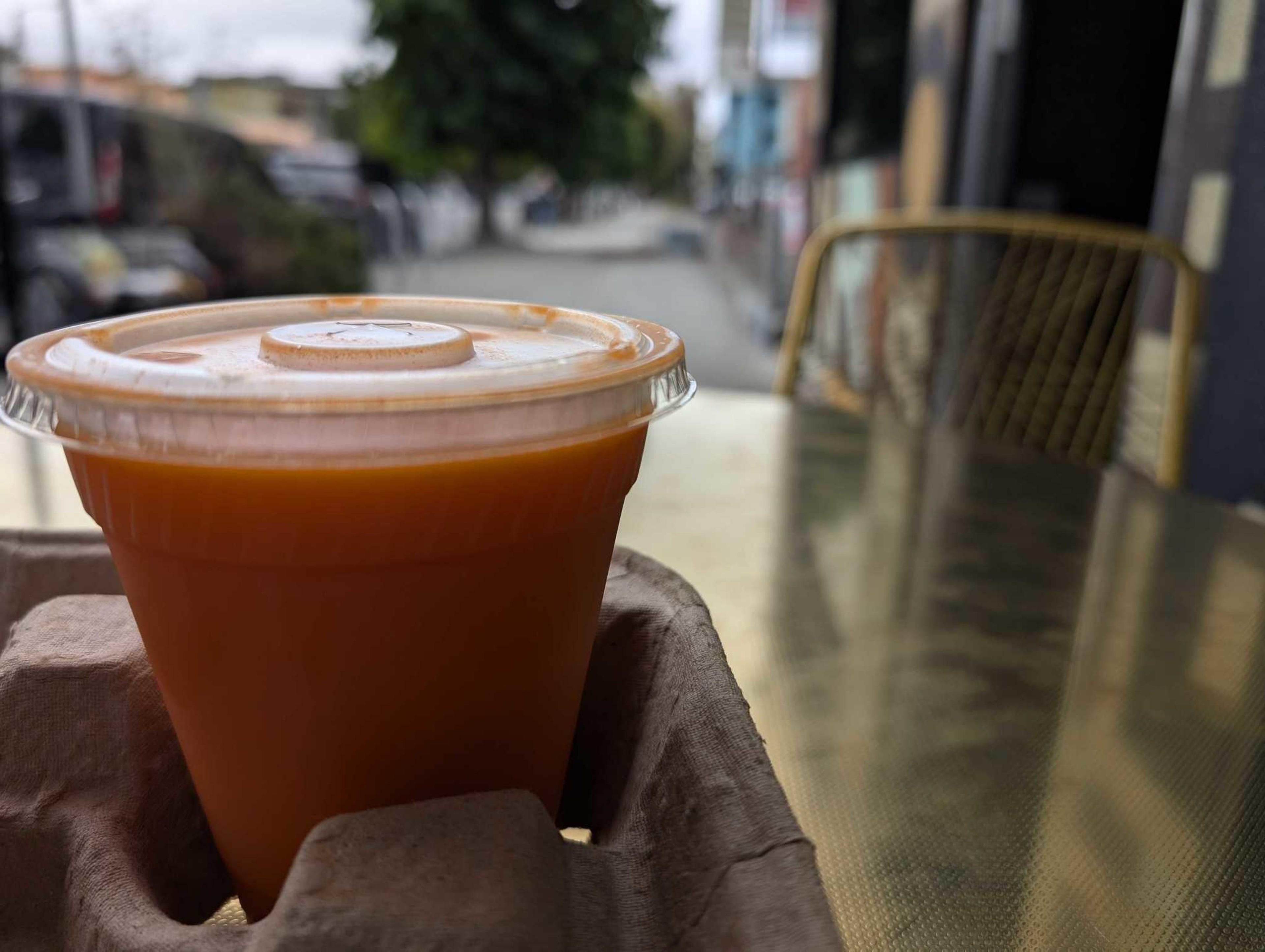 A plastic cup filled with orange juice sits on a cardboard holder on a metal table, with a blurred outdoor street view in the background.
