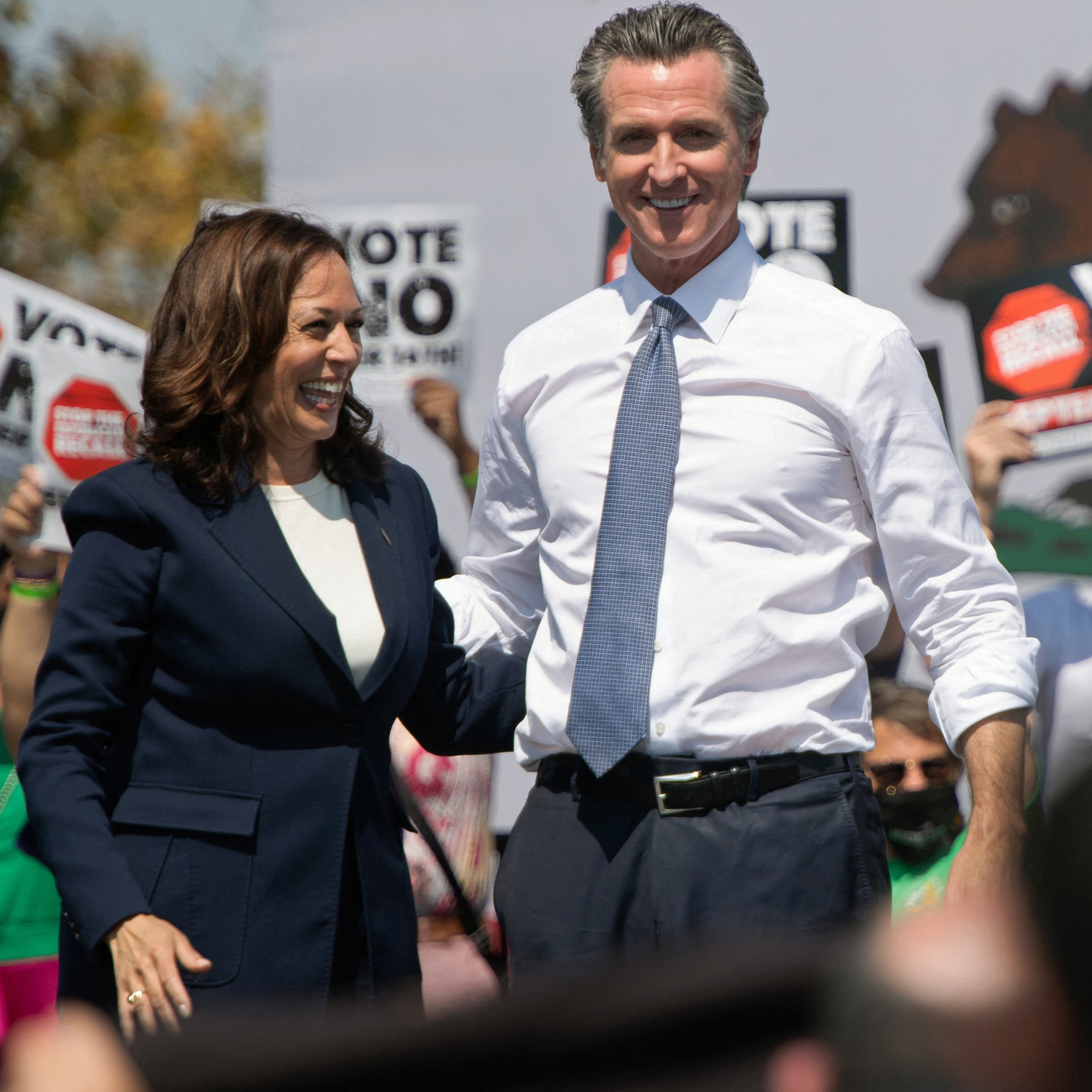 A man in a white shirt and tie with rolled-up sleeves stands beside a woman in a navy blazer. They are smiling amid a crowd holding &quot;Vote No&quot; signs.
