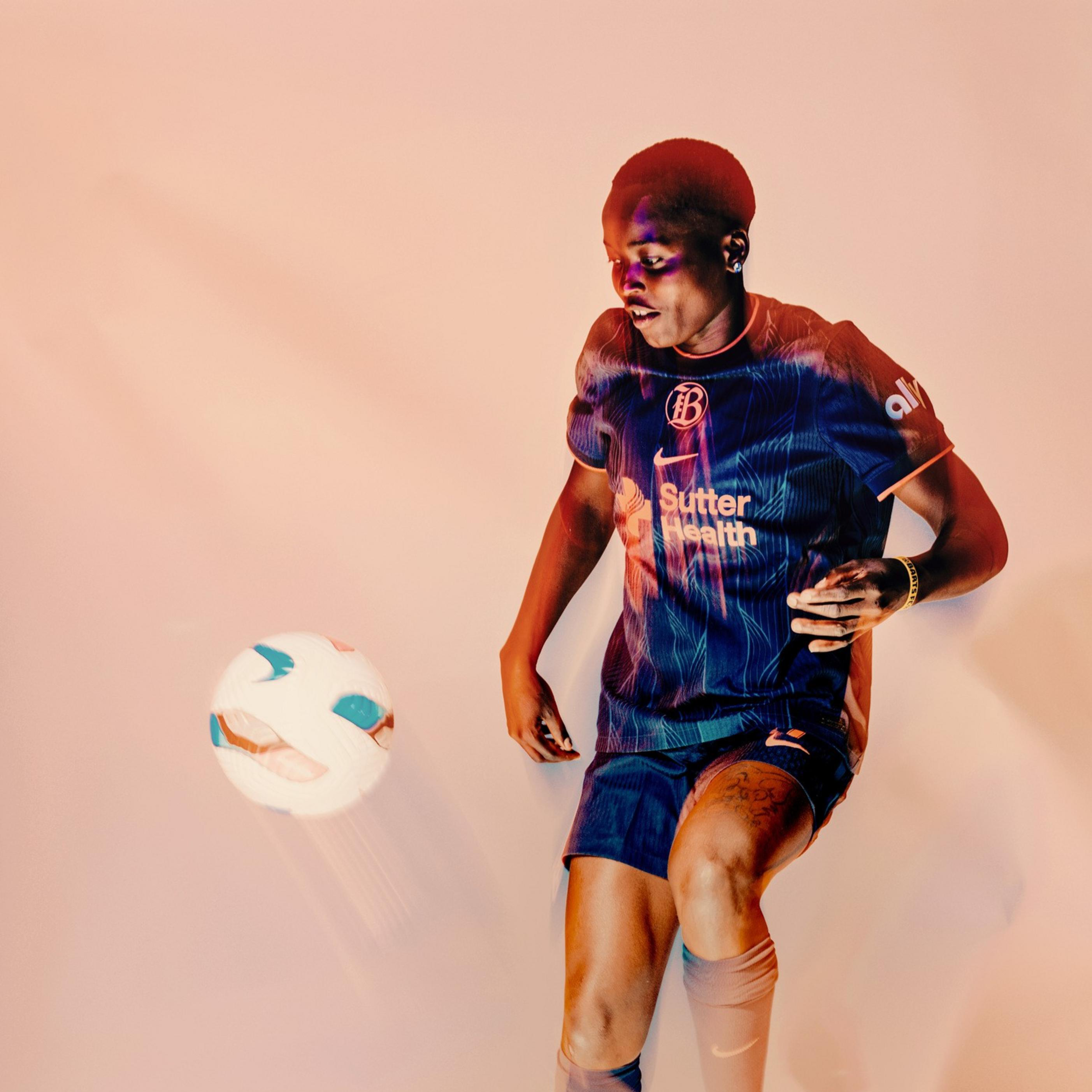 A soccer player in a blue uniform is mid-action, controlling a white and blue ball with their left knee against a plain, light background.