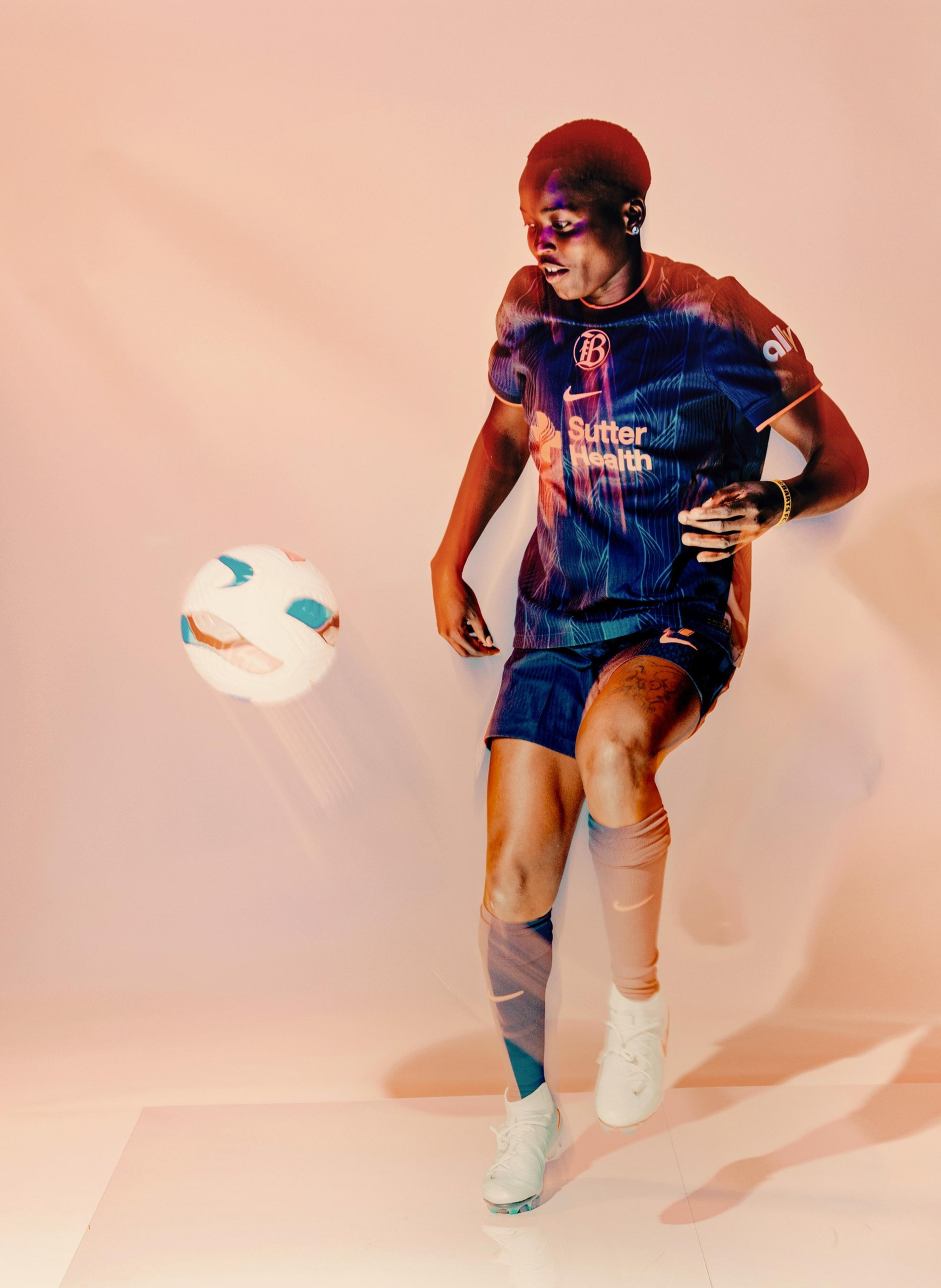 A soccer player in a blue uniform is mid-action, controlling a white and blue ball with their left knee against a plain, light background.