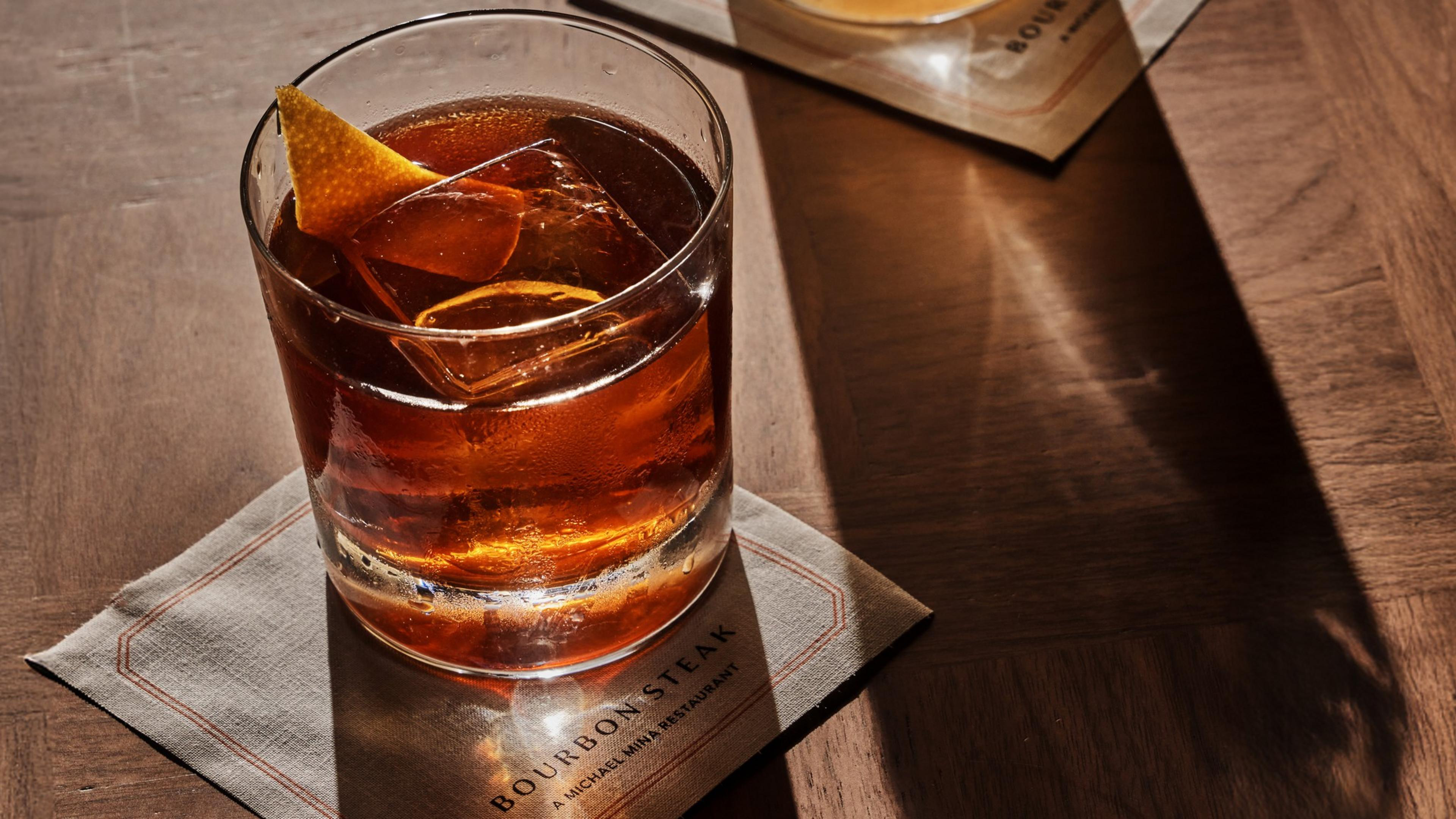A glass of dark amber cocktail with a large ice cube and an orange peel garnish rests on a Bourbon Steak napkin on a wooden table.