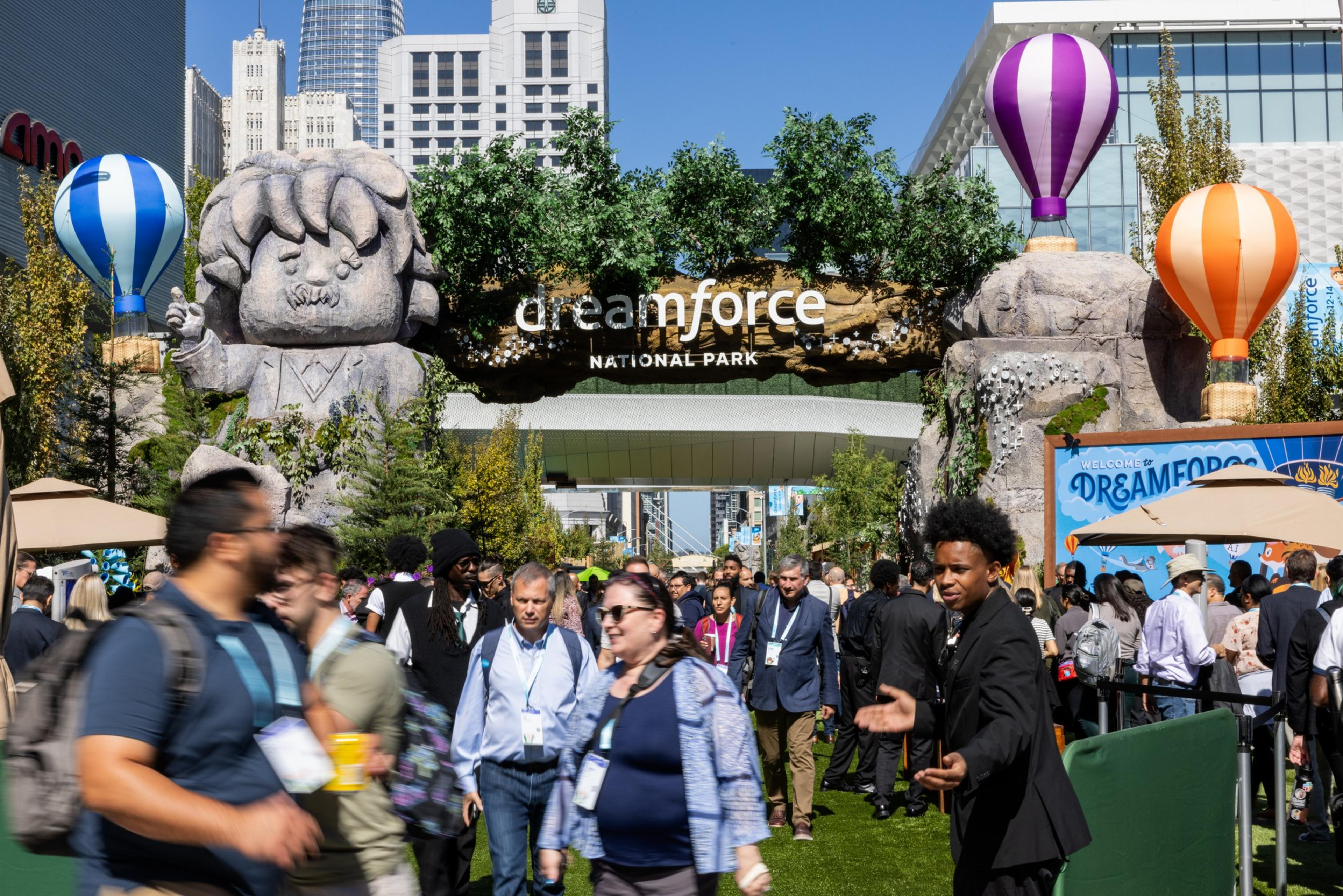 The image shows a crowded event entrance labeled &quot;Dreamforce National Park,&quot; decorated with large, cartoonish statues, colorful hot air balloons, and green foliage under a clear sky.
