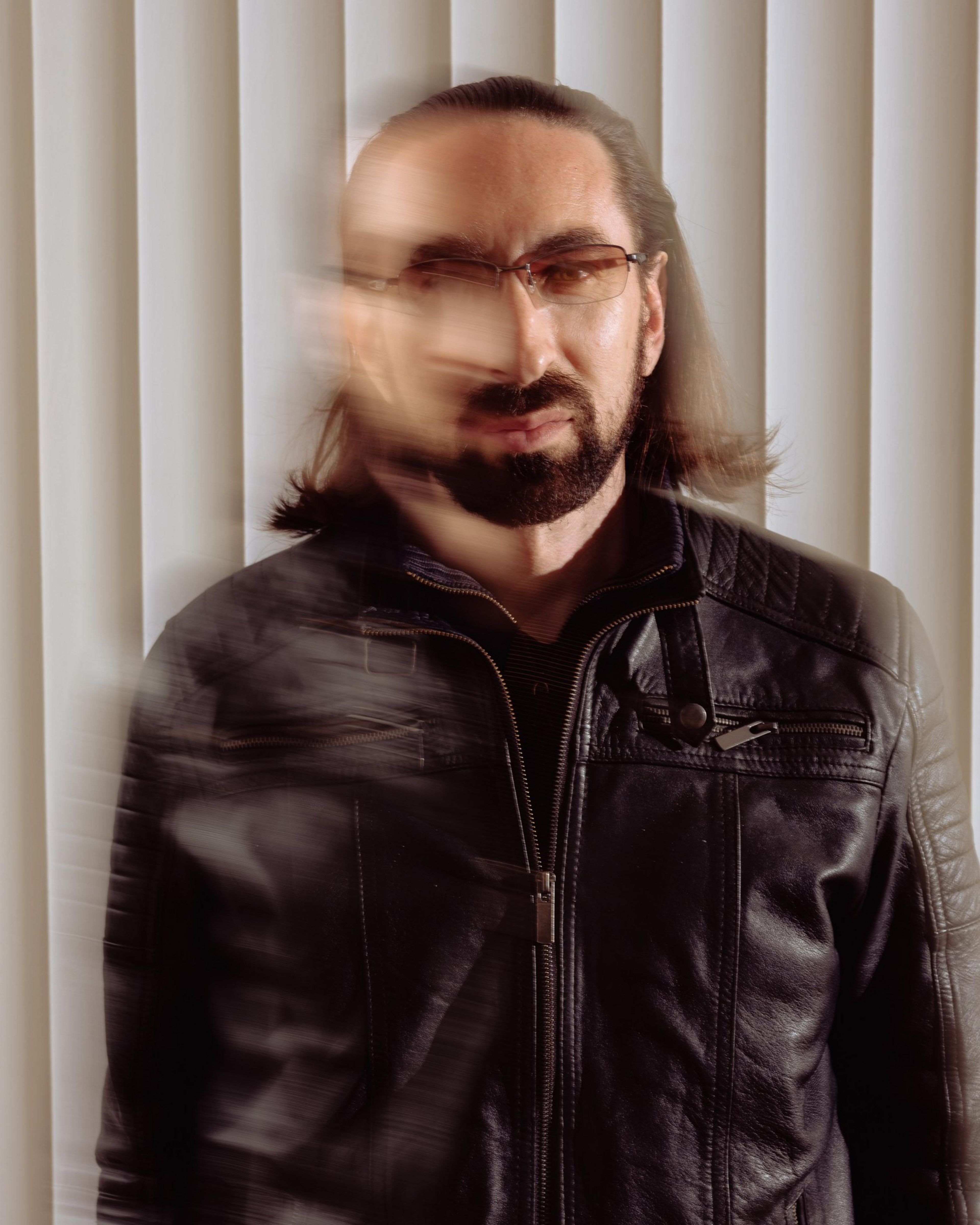 A man with long hair and glasses wears a black leather jacket. The image features a blurred effect, creating a sense of movement across his face and torso.