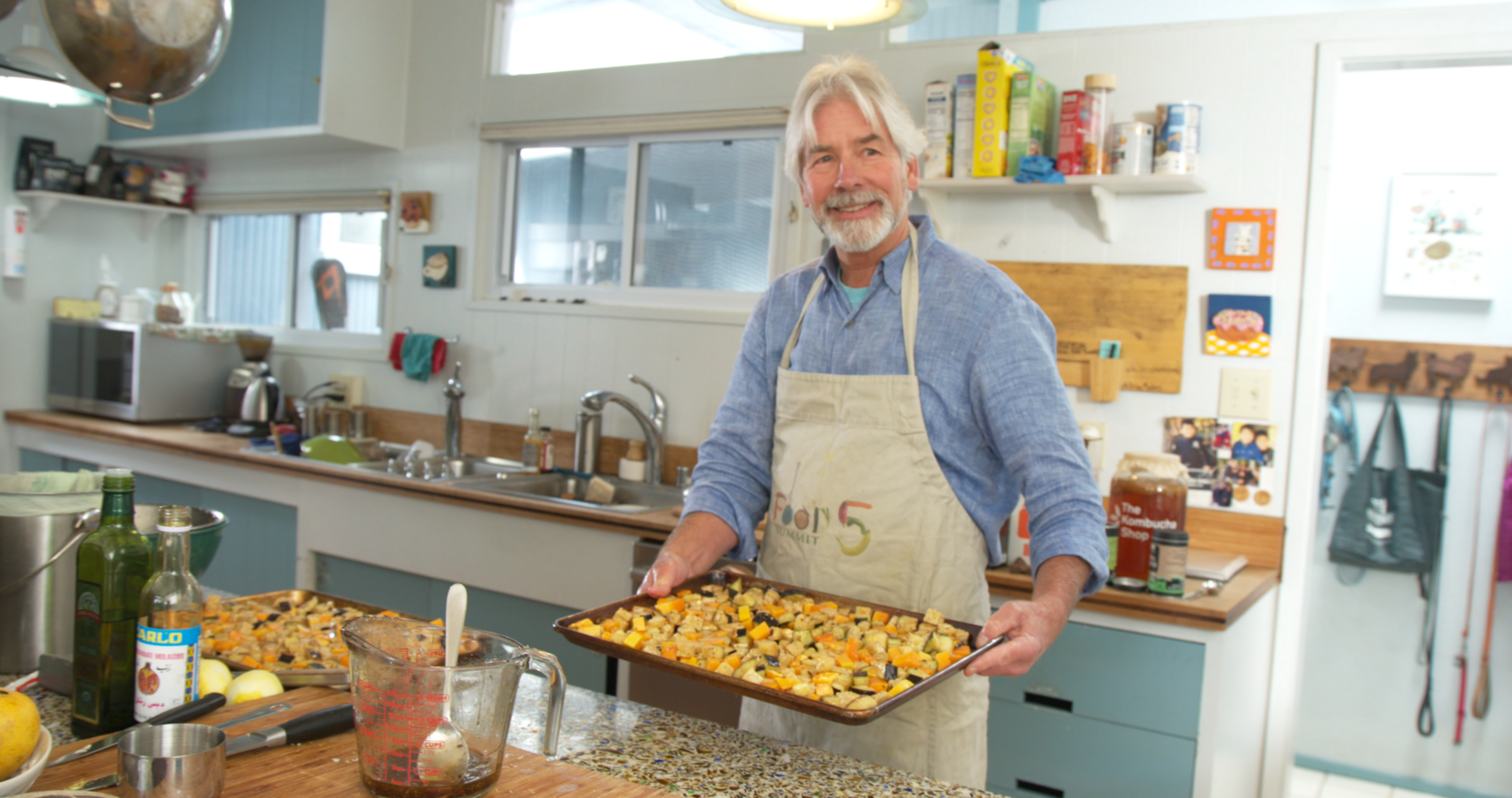 A man stands in a kitchen holding a tray of eggplant and tempeh.