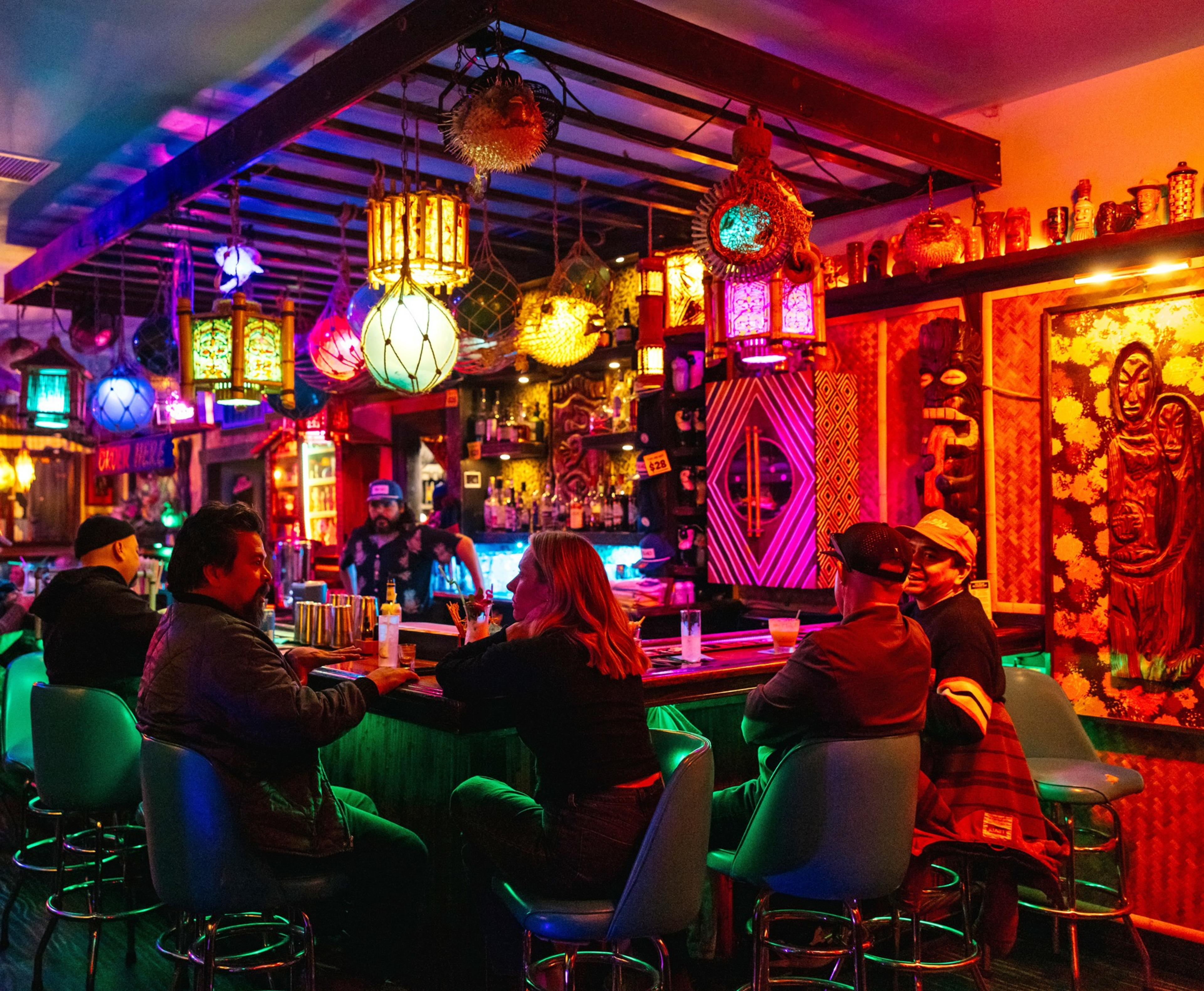 A vibrant bar with colorful hanging lanterns and eclectic decor. Five people sit at the bar, engaging in conversation under a warm, glowing atmosphere.