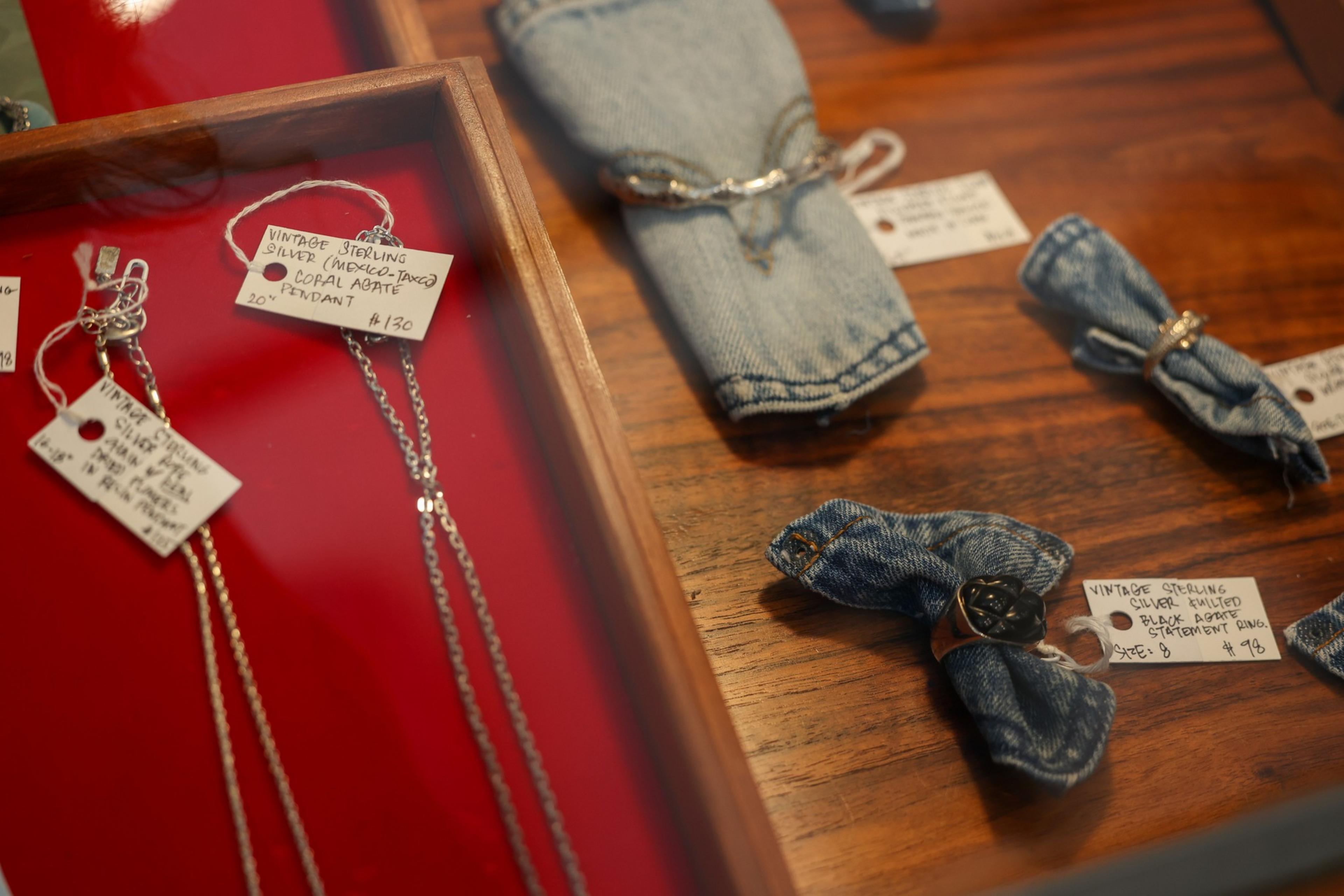Vintage sterling silver jewelry, including necklaces with pendants and rings displayed on denim fabric and wooden surfaces, each with handwritten price tags.