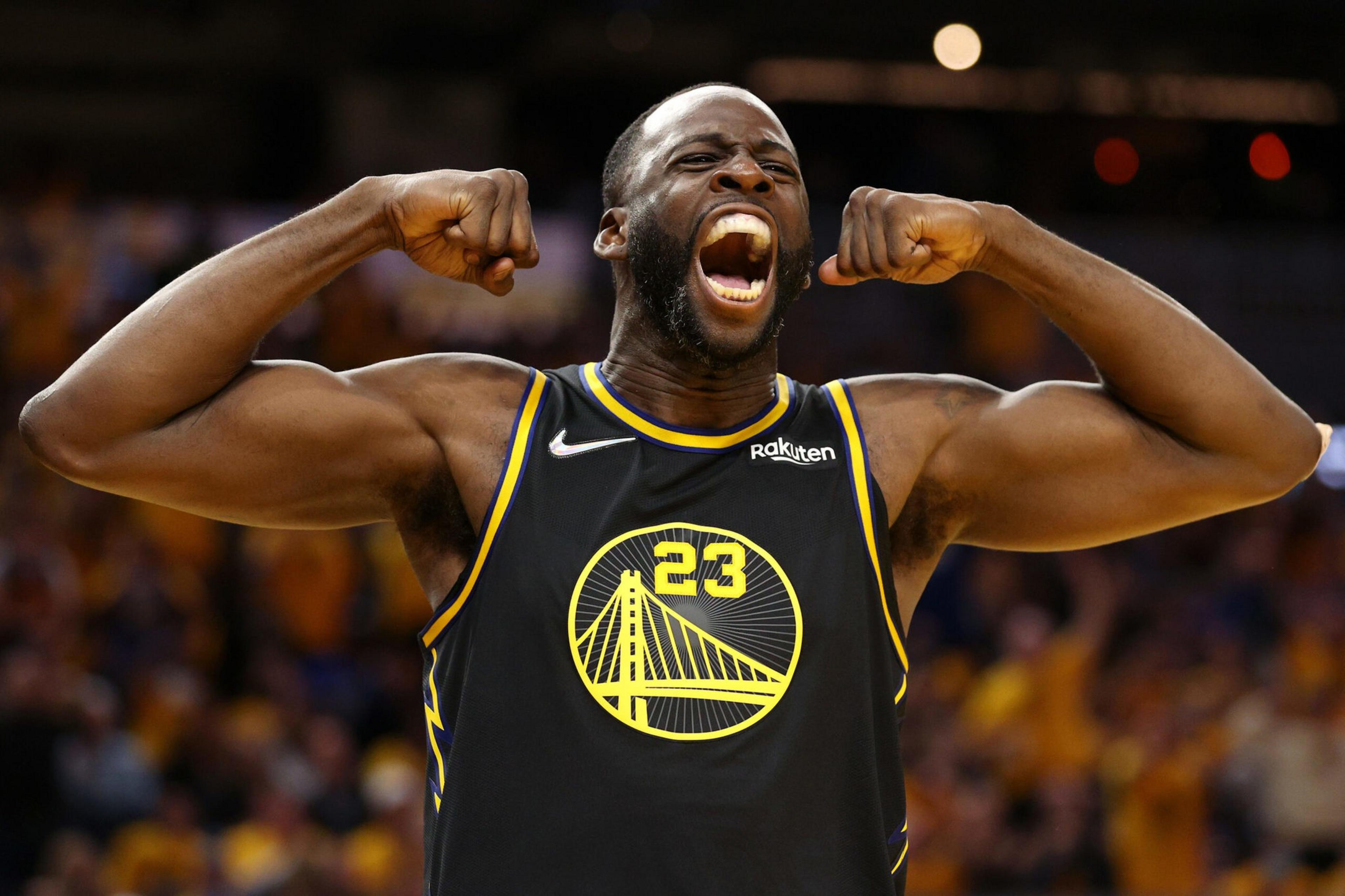 The image shows a basketball player wearing a black jersey with yellow accents, number 23. He's flexing his arms and shouting energetically, with a blurred crowd in the background.