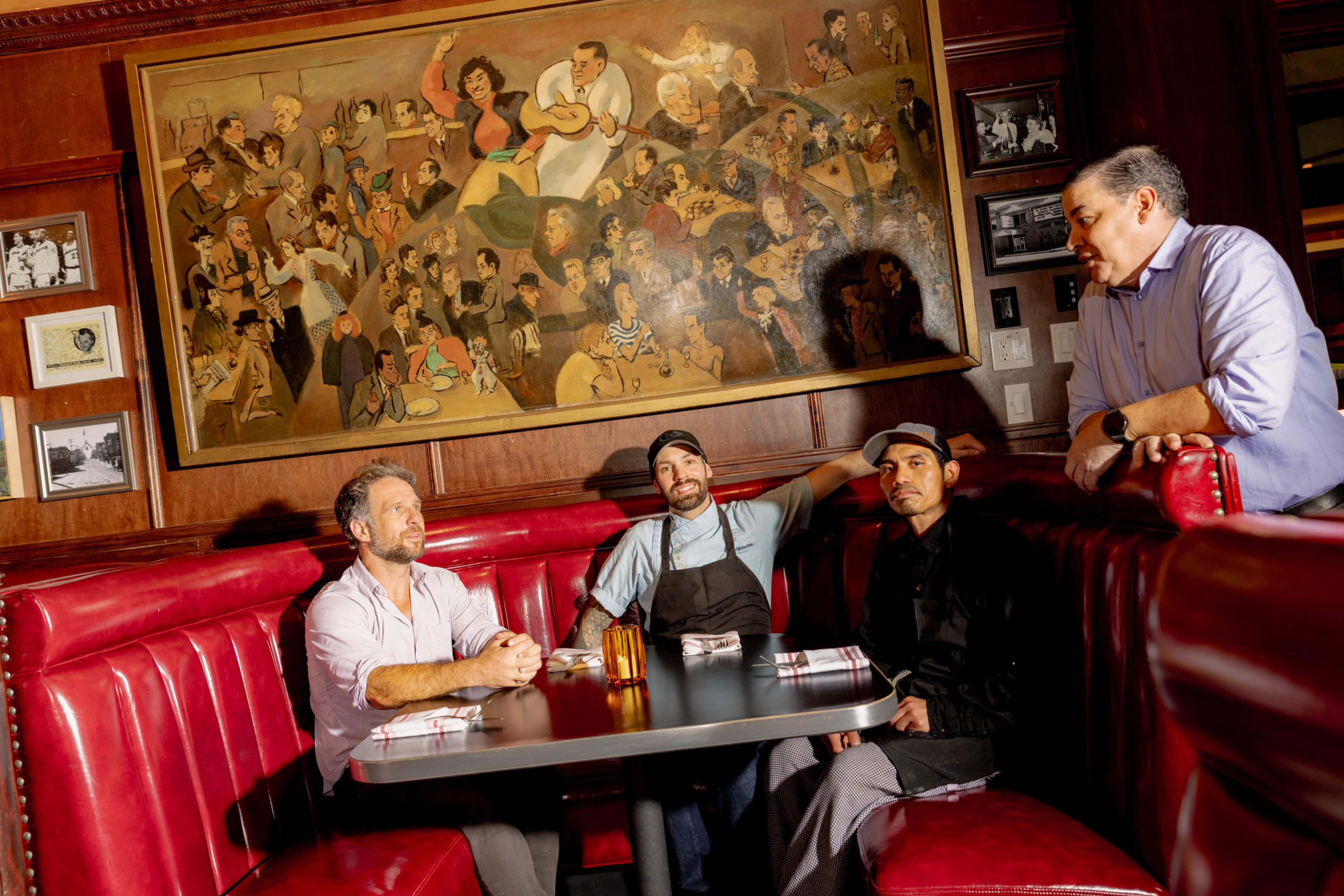 Four men sit and lean around a red leather booth in a restaurant, with a large colorful painting of people playing music and socializing hanging on the wall behind them.